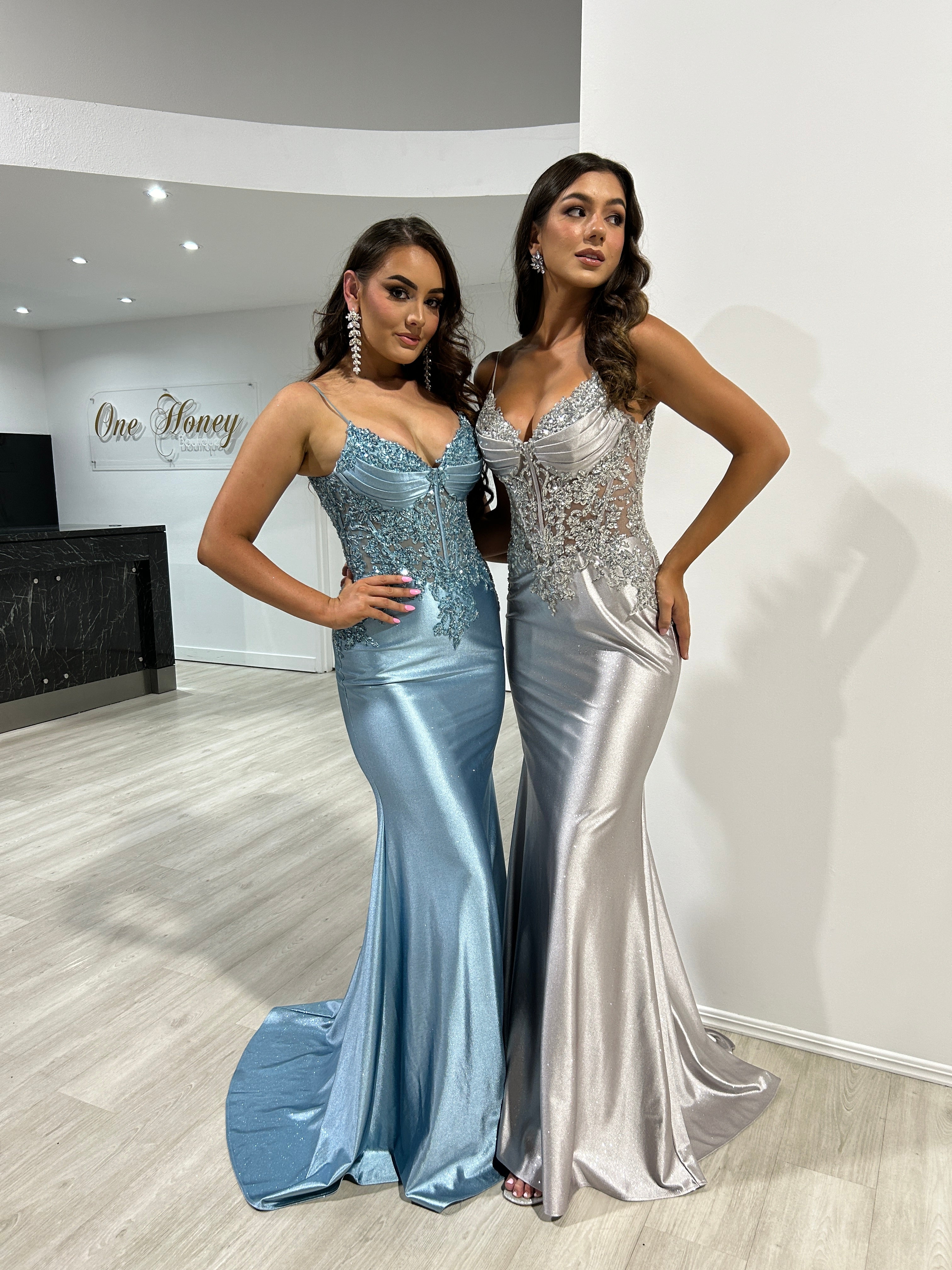 Two women wearing elegant silver and blue satin mermaid prom dresses with beaded bodices, standing indoors. One Honey, Brisbane to Gold Coast.