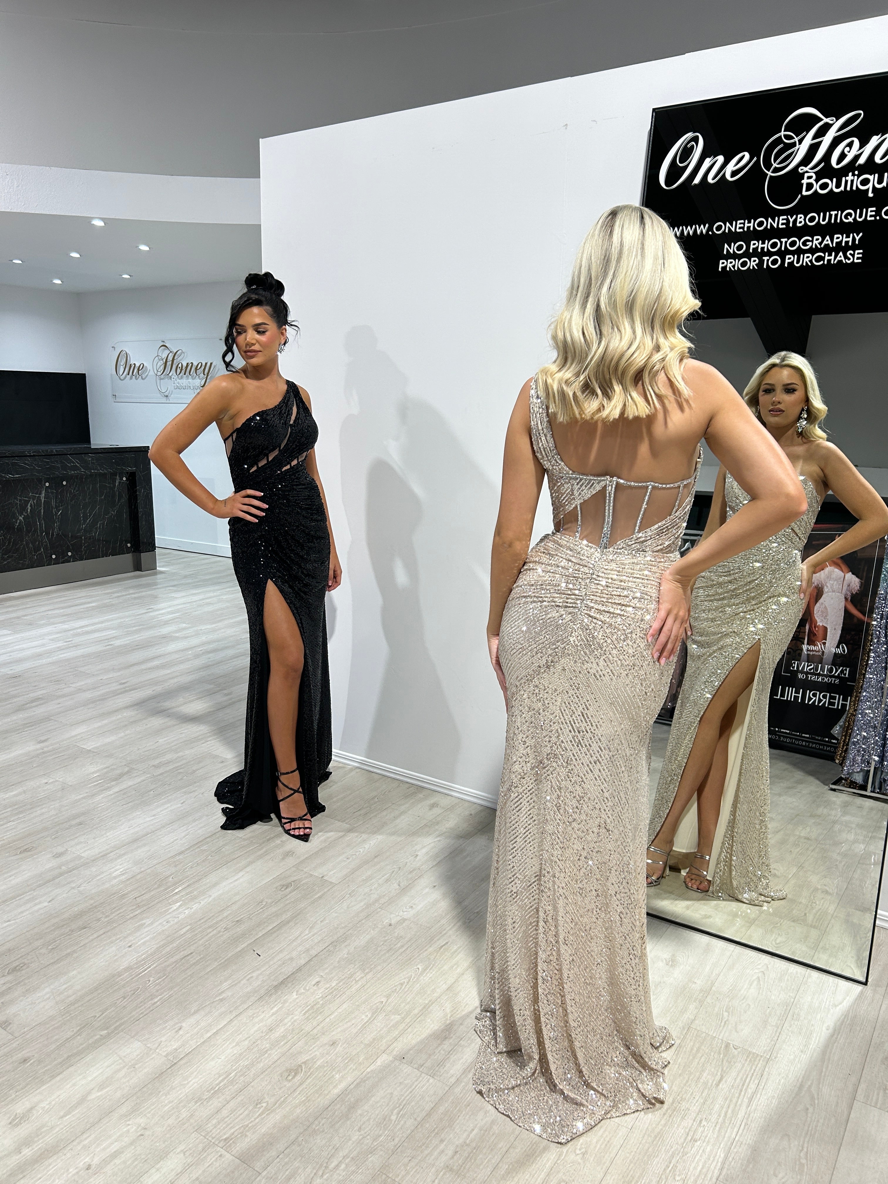 Two women model dresses from Honey Couture, one in a black gown and the other in IRIS Platinum Silver One Shoulder Sequin Corset Formal Dress.