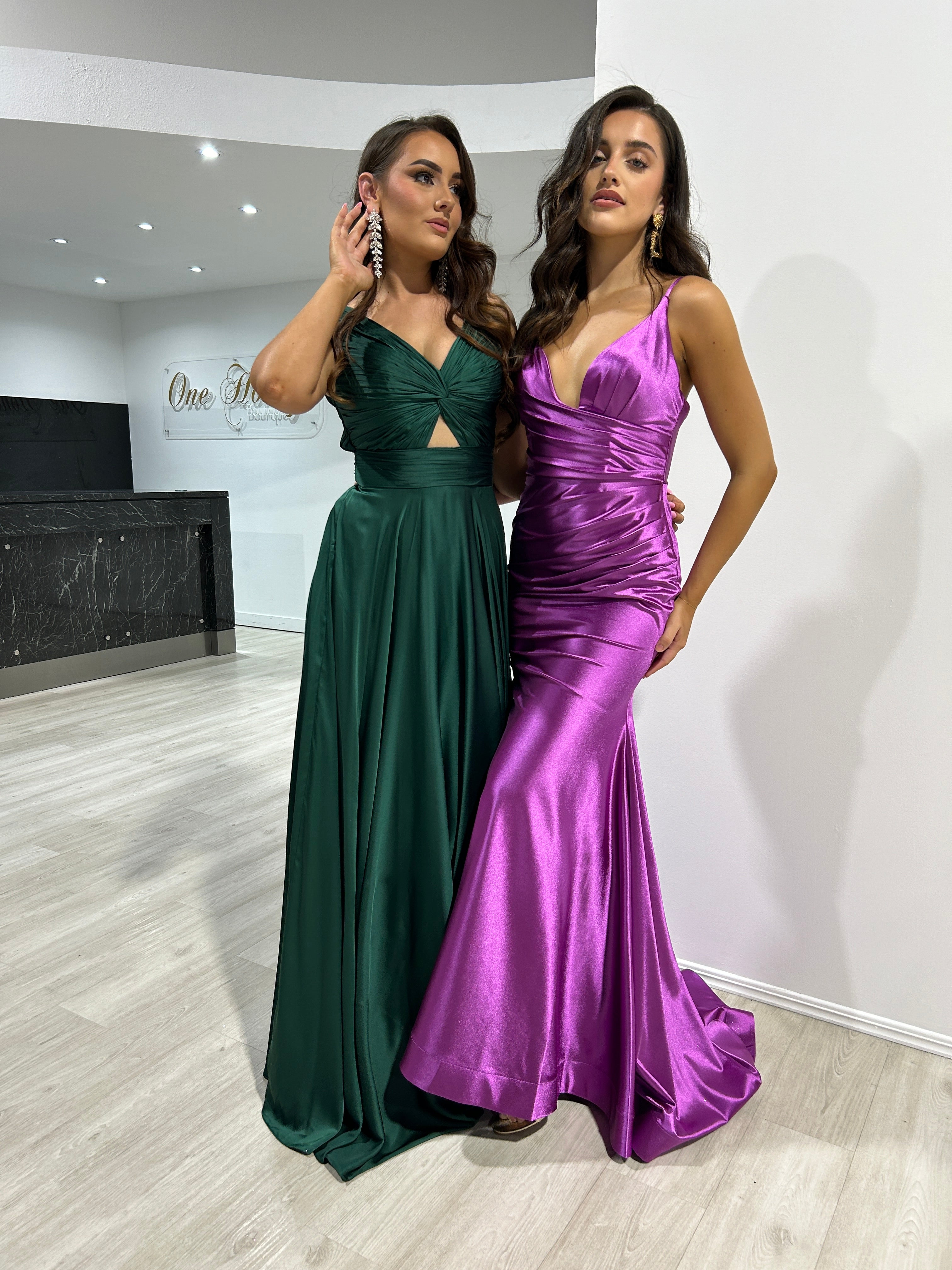 Two women wearing elegant formal dresses, one in an emerald green Honey Couture WILLOWMENA A-line, posing in a stylish interior.