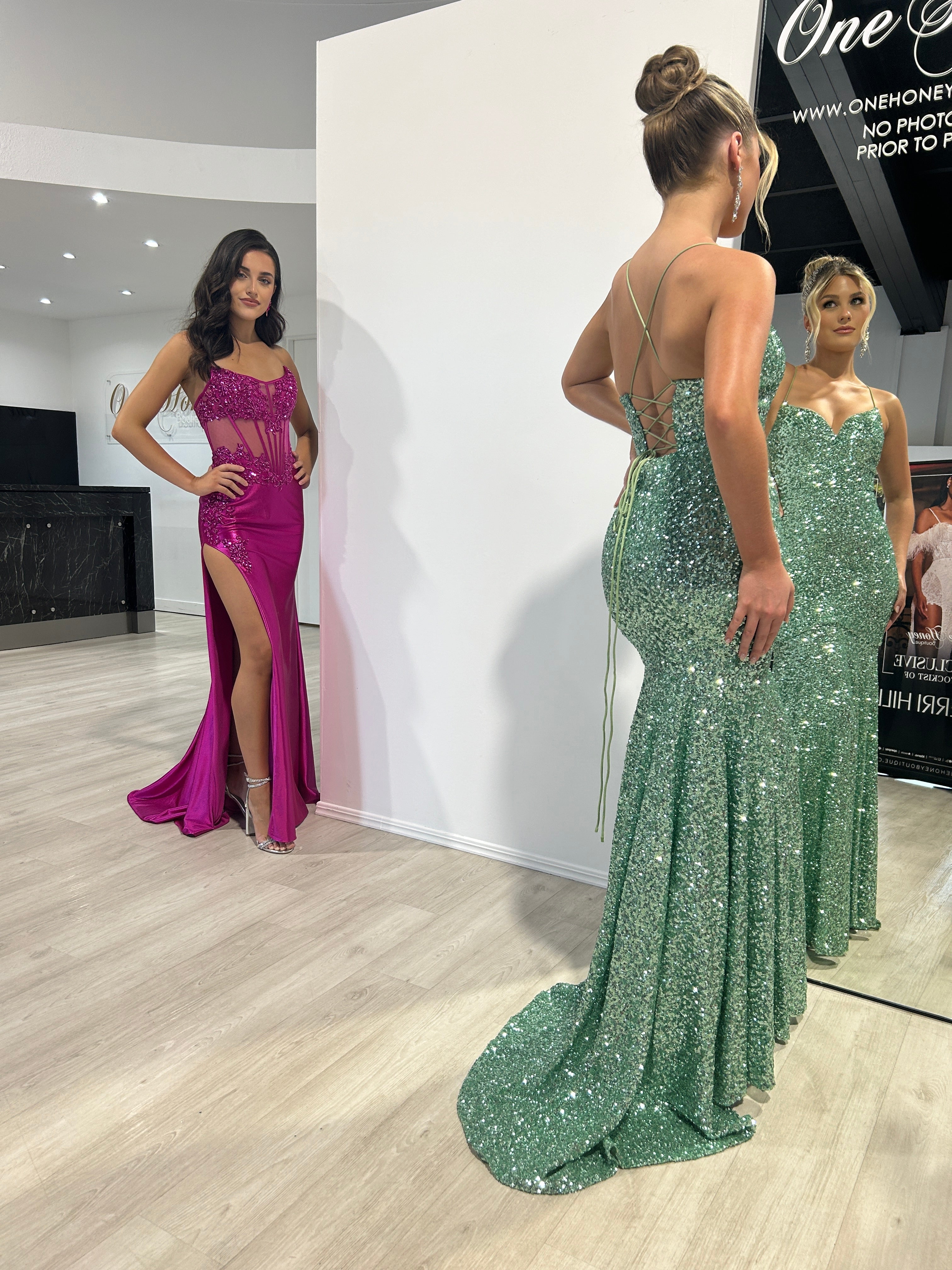 A woman in a green glittered backless gown with a strappy design stands in a boutique, admiring her reflection, while another woman in a purple dress stands nearby.