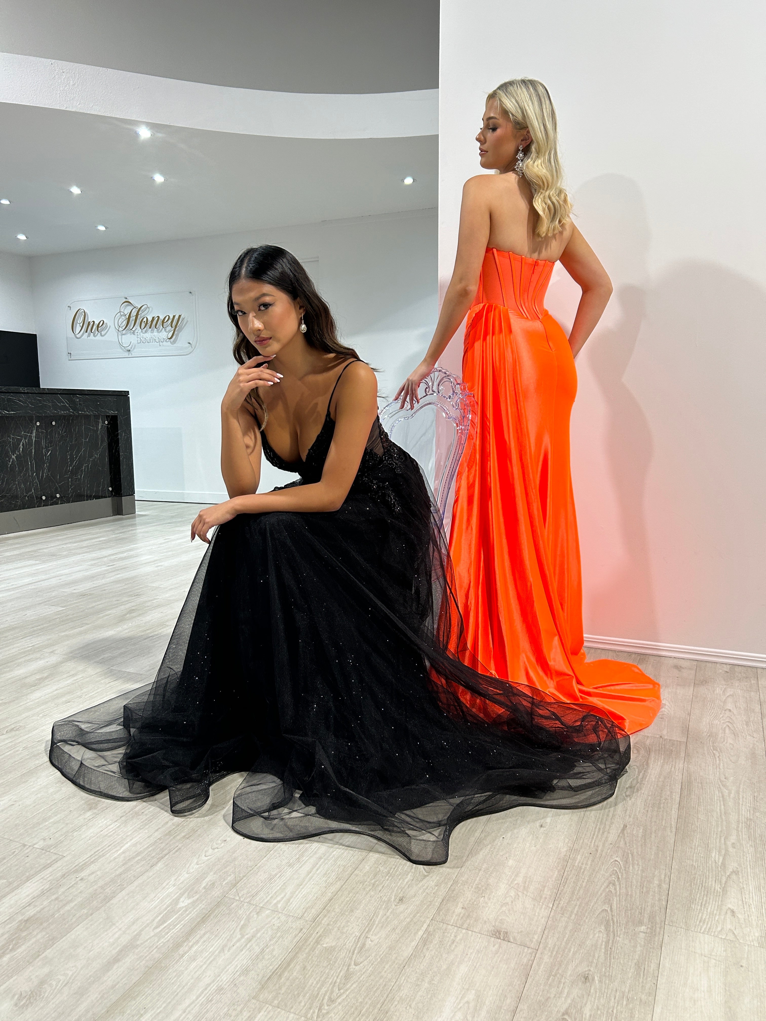 Two women in formal dresses, one in a black tulle evening gown and the other in a striking orange strapless dress, posing elegantly indoors.