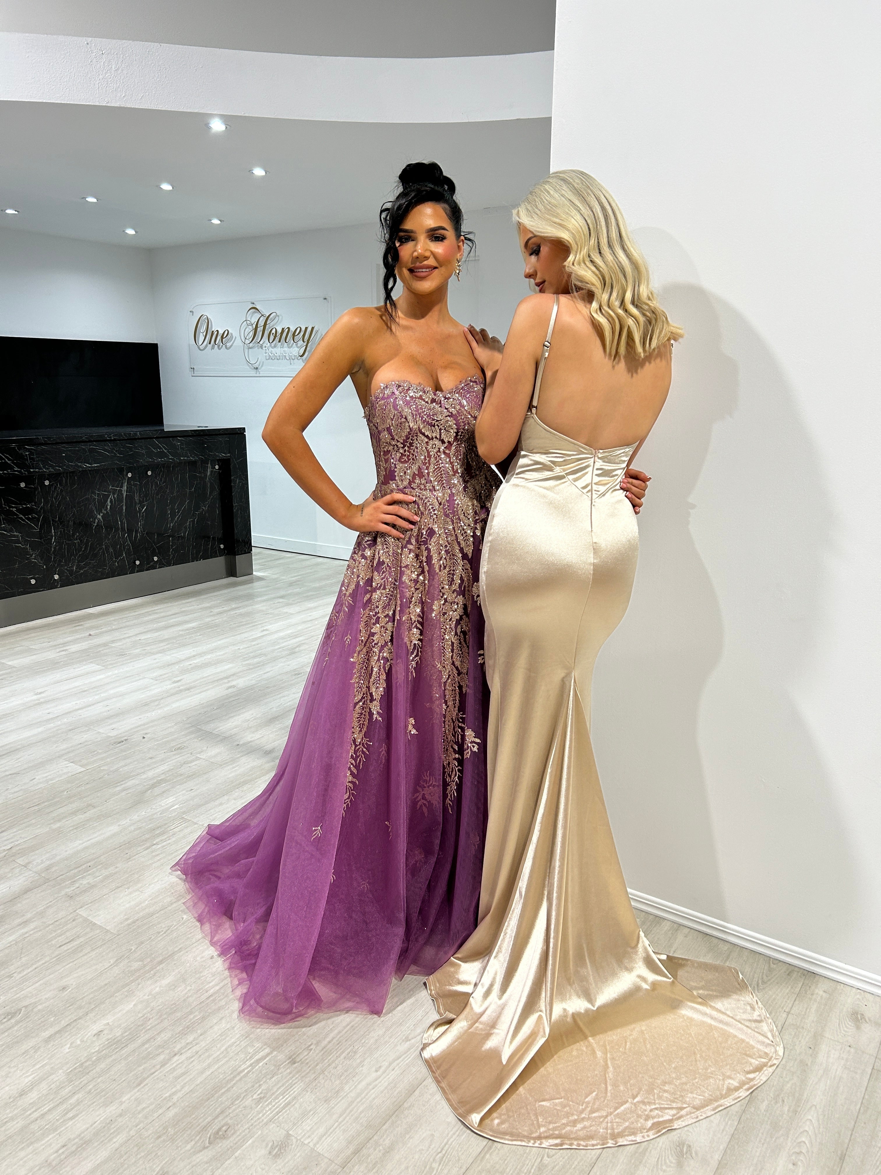 Two women in elegant evening gowns; one in a purple gown with lace embellishments, the other in a champagne satin dress.