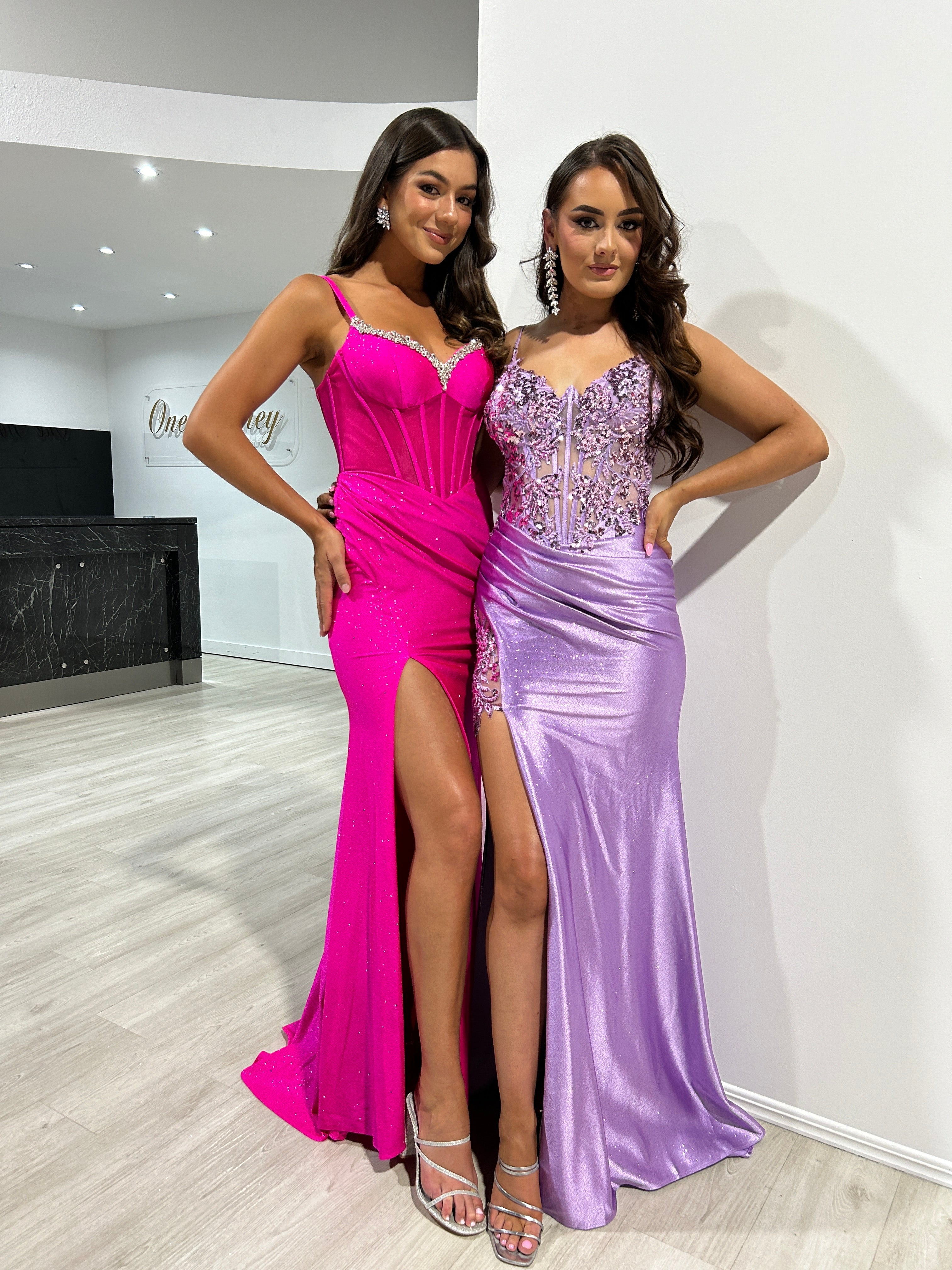 Two women wearing glamorous evening gowns with glitter corset details and high slits, standing side by side indoors.