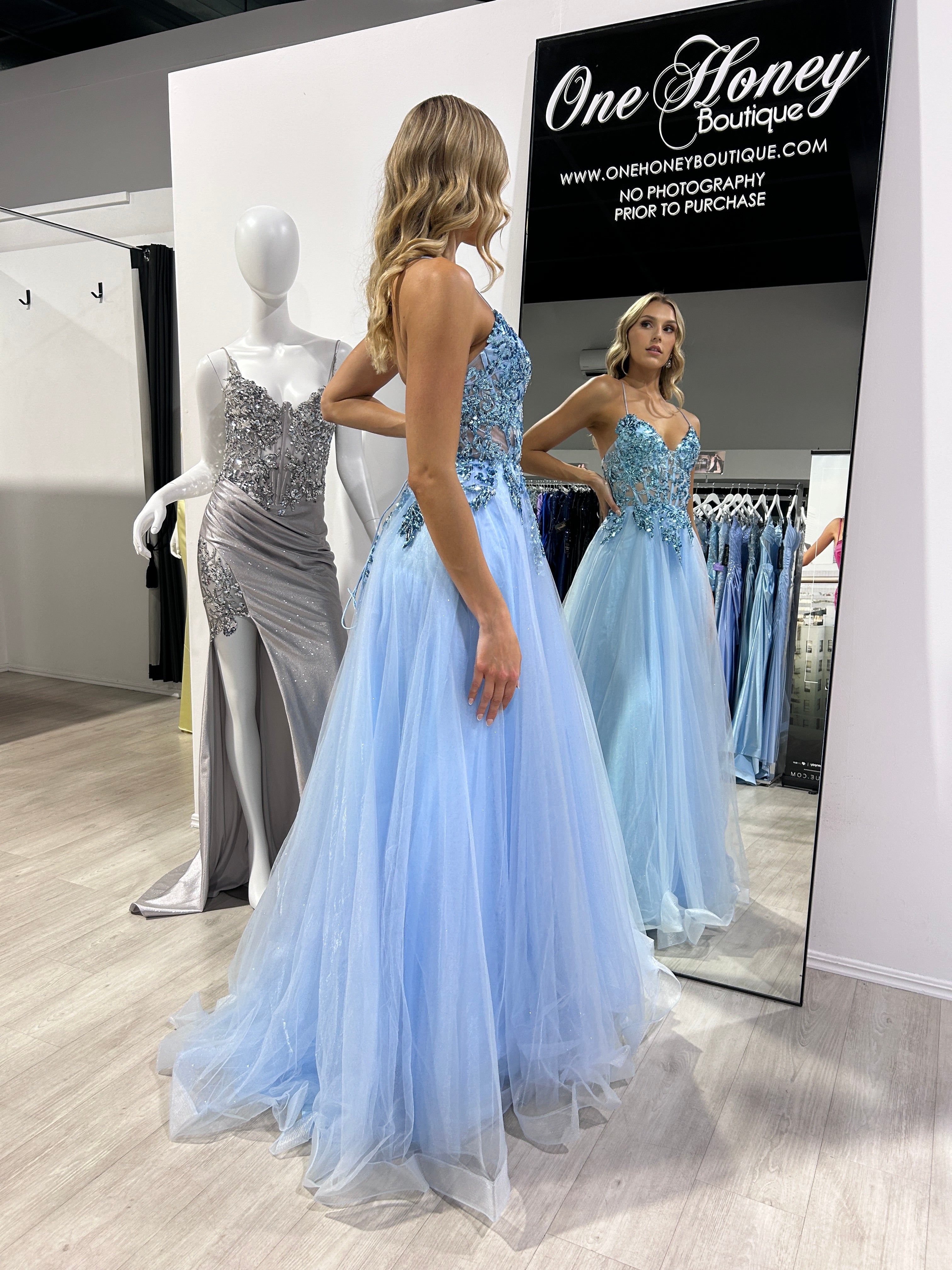 Woman in blue formal dress with lace and tulle admires reflection, surrounded by dresses, One Honey Boutique.