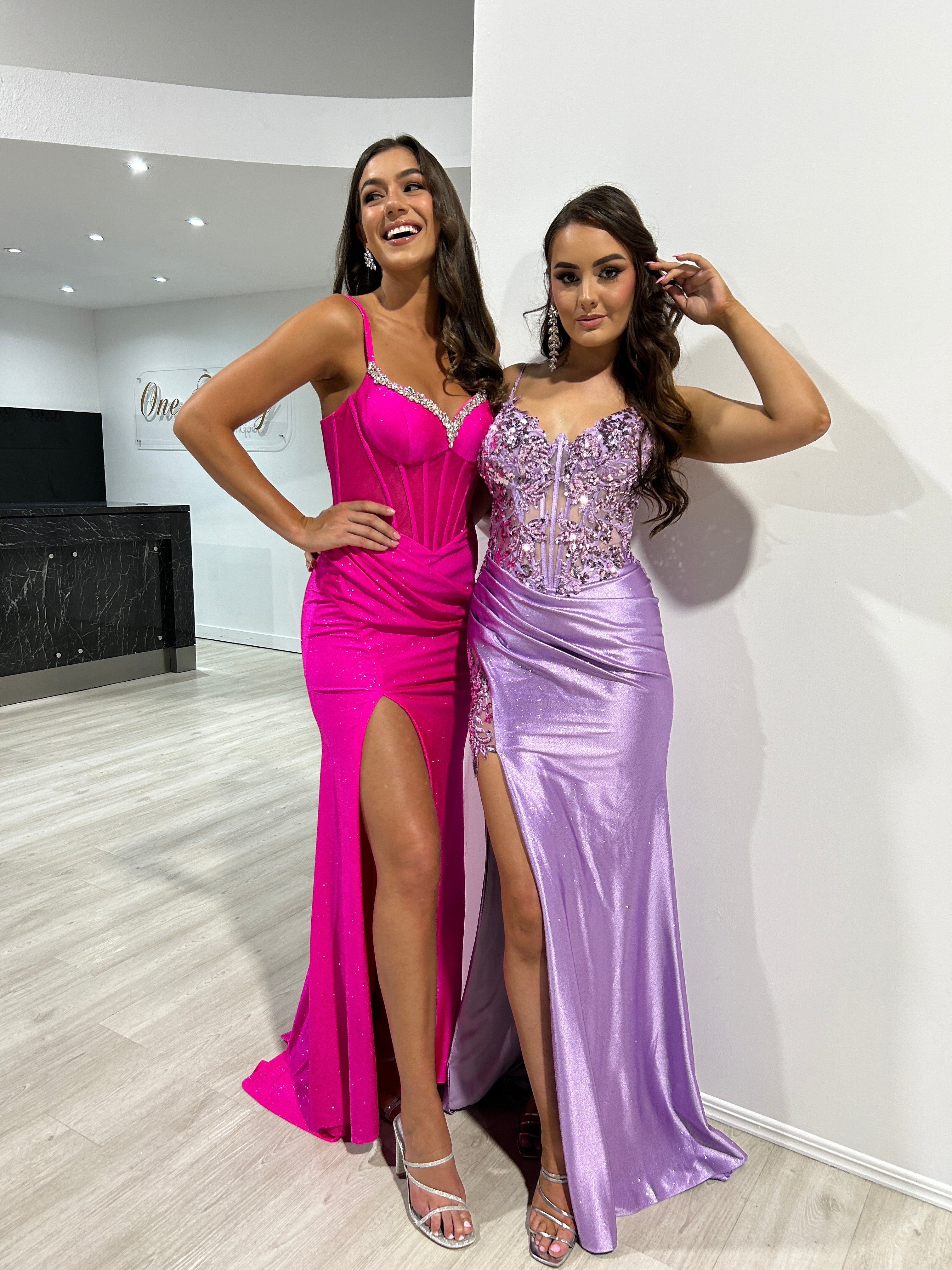 Two women in glittering evening gowns, one in vibrant pink with a side slit and the other in lilac with intricate beading, posing indoors.