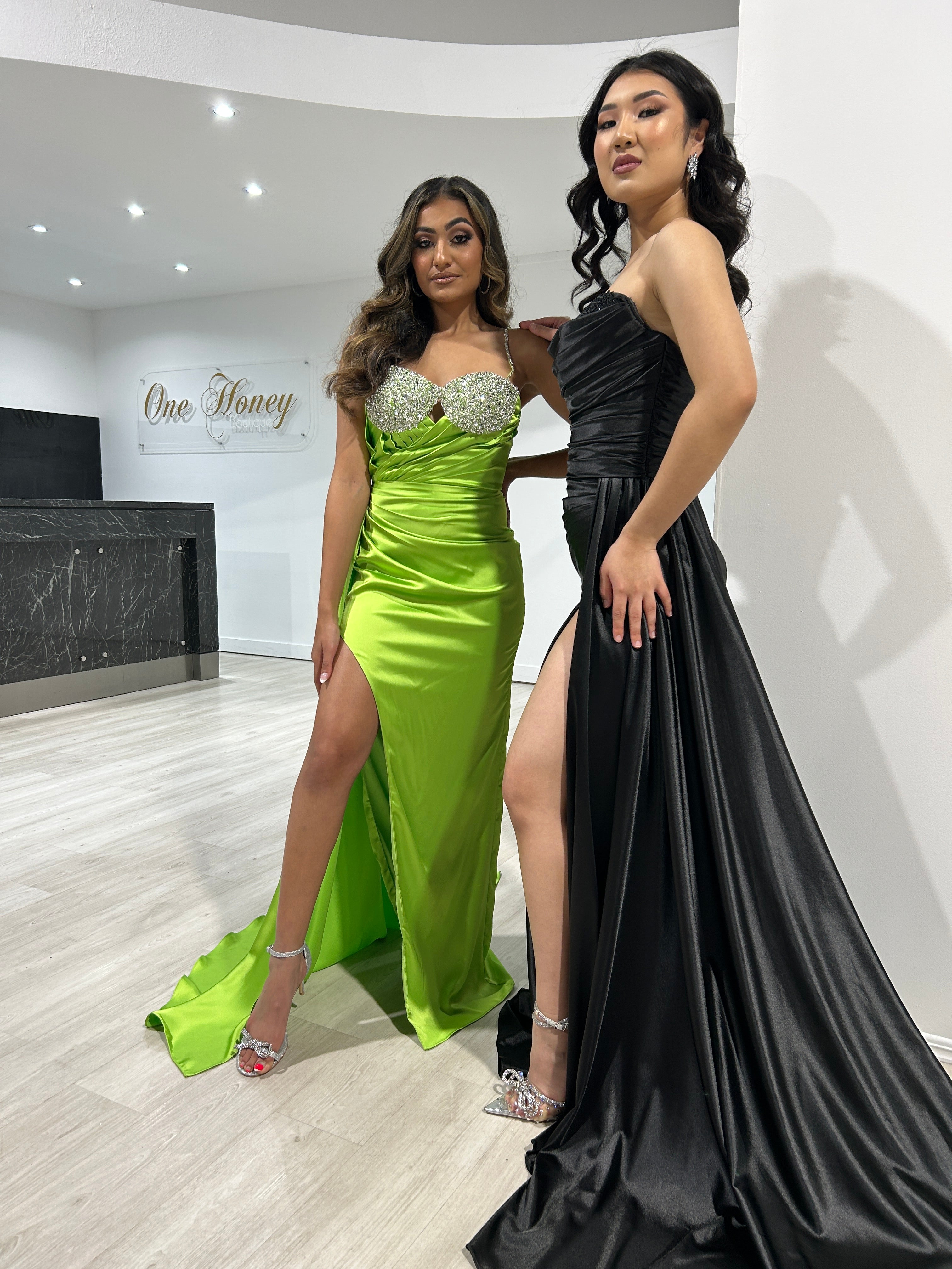 Two women in bright green and black formal gowns with leg splits, posing in a modern room with a sign reading 'One Honey'.