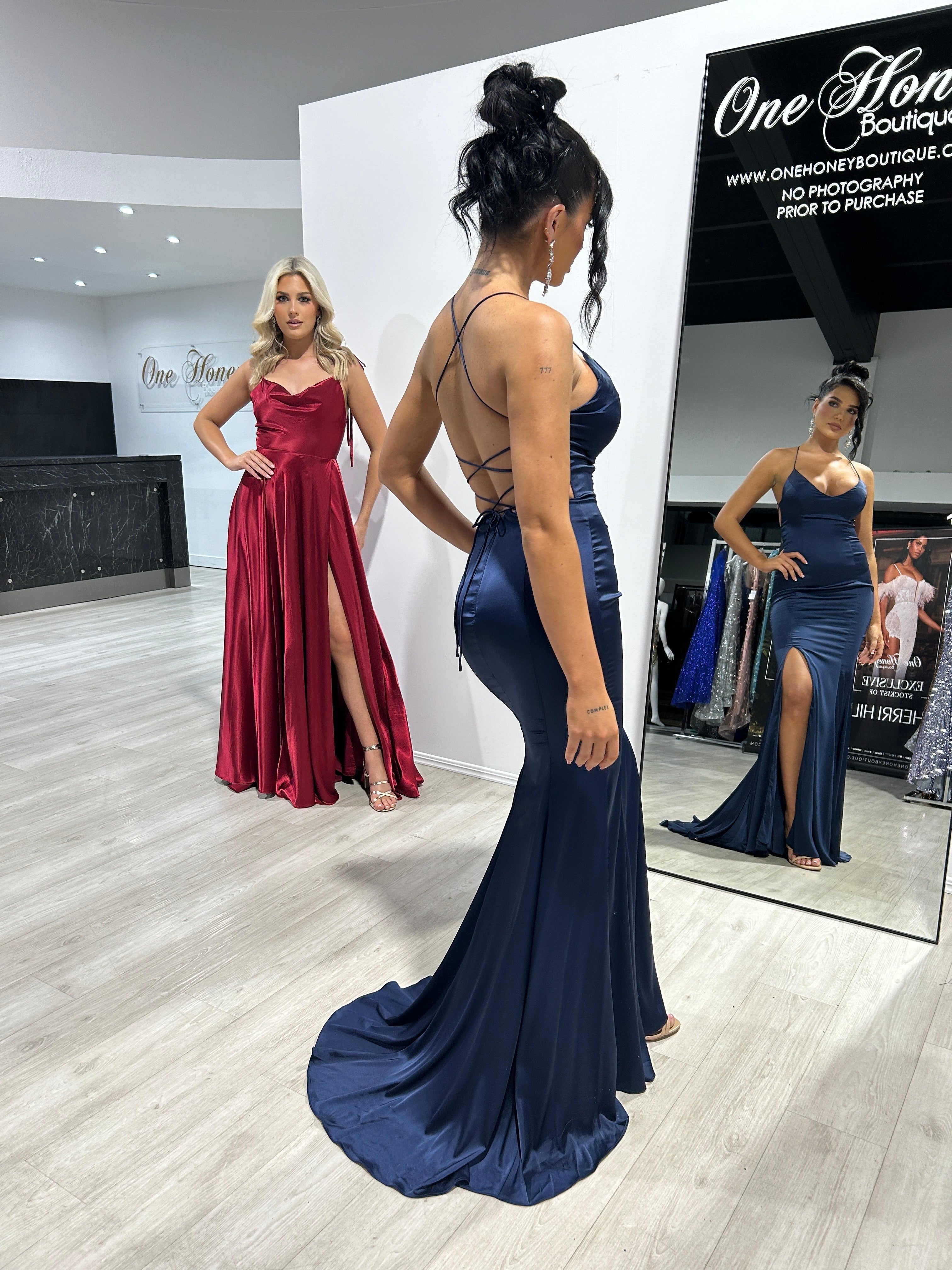 Two women model elegant formal gowns with spaghetti straps and slits on a boutique floor, showcasing flowing silhouettes in red and navy.
