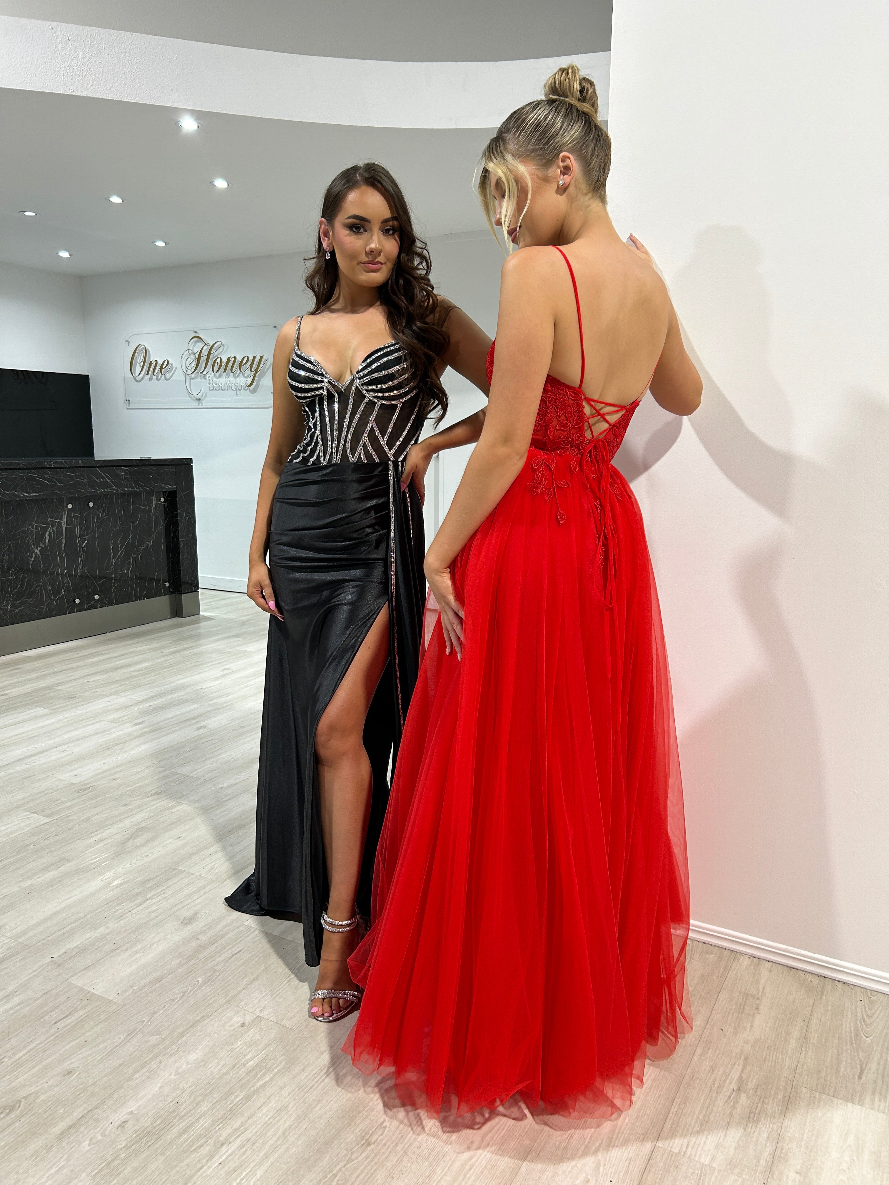 Two women in formal dresses; one in a black gown and another in Honey Couture JARRISSA red tulle dress with lace applique.