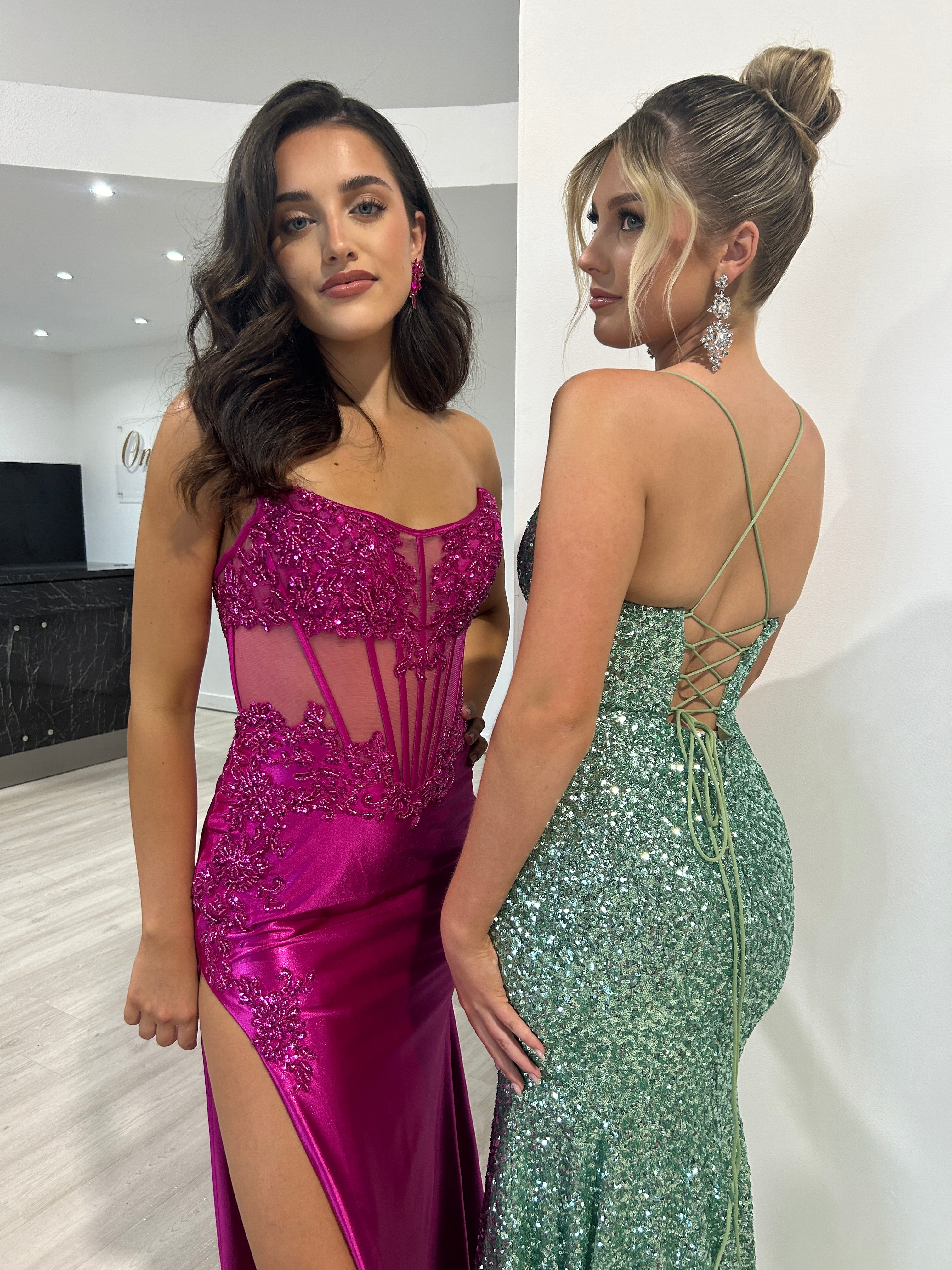 Two women in elegant evening gowns; one wears a magenta sheer corset satin dress, the other a green sequin backless dress. Brisbane to Gold Coast.