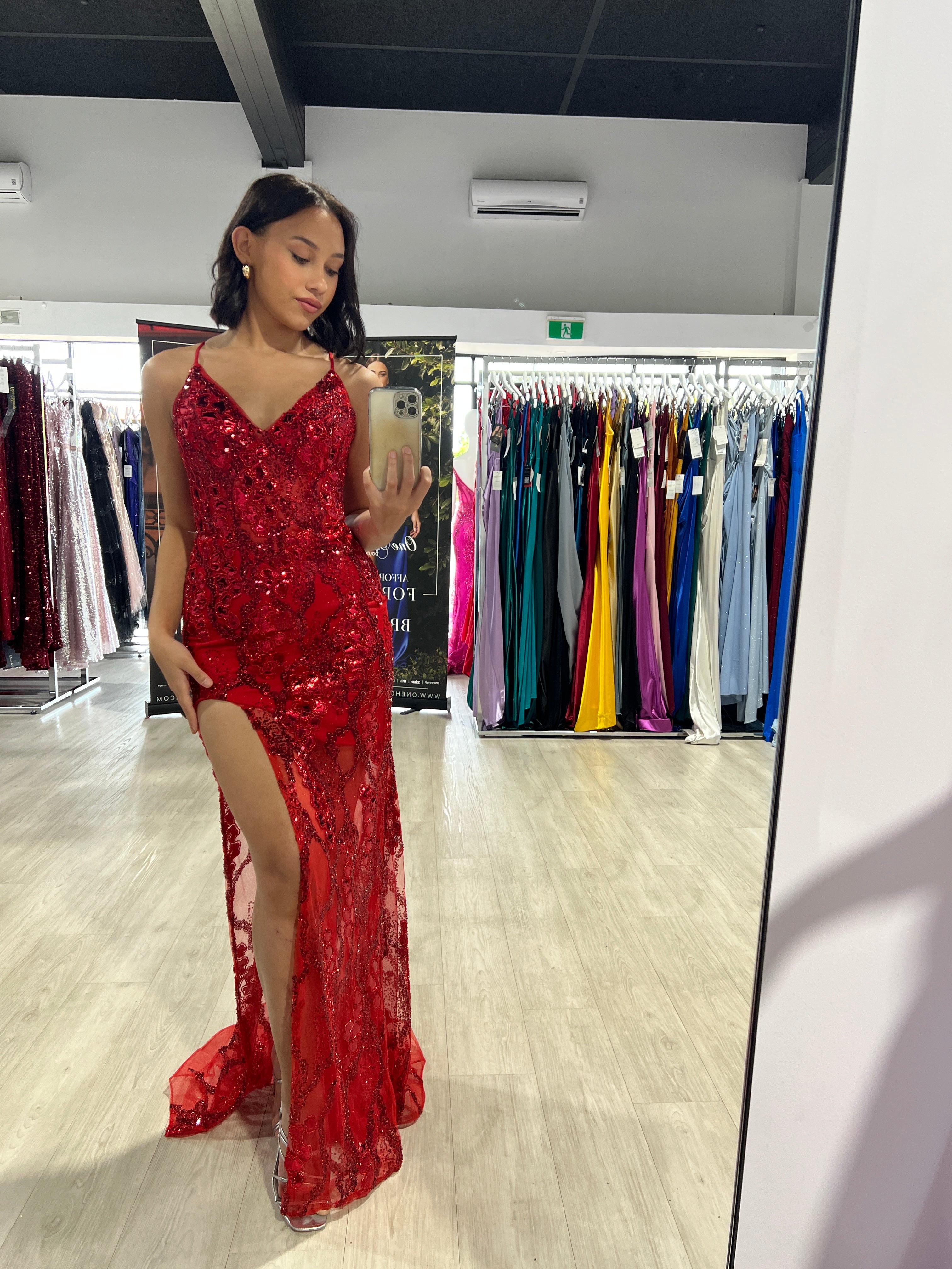 Elegant red sequin evening gown with sheer mesh skirt and side slit, displayed in a boutique. Mirror reflection adds depth.