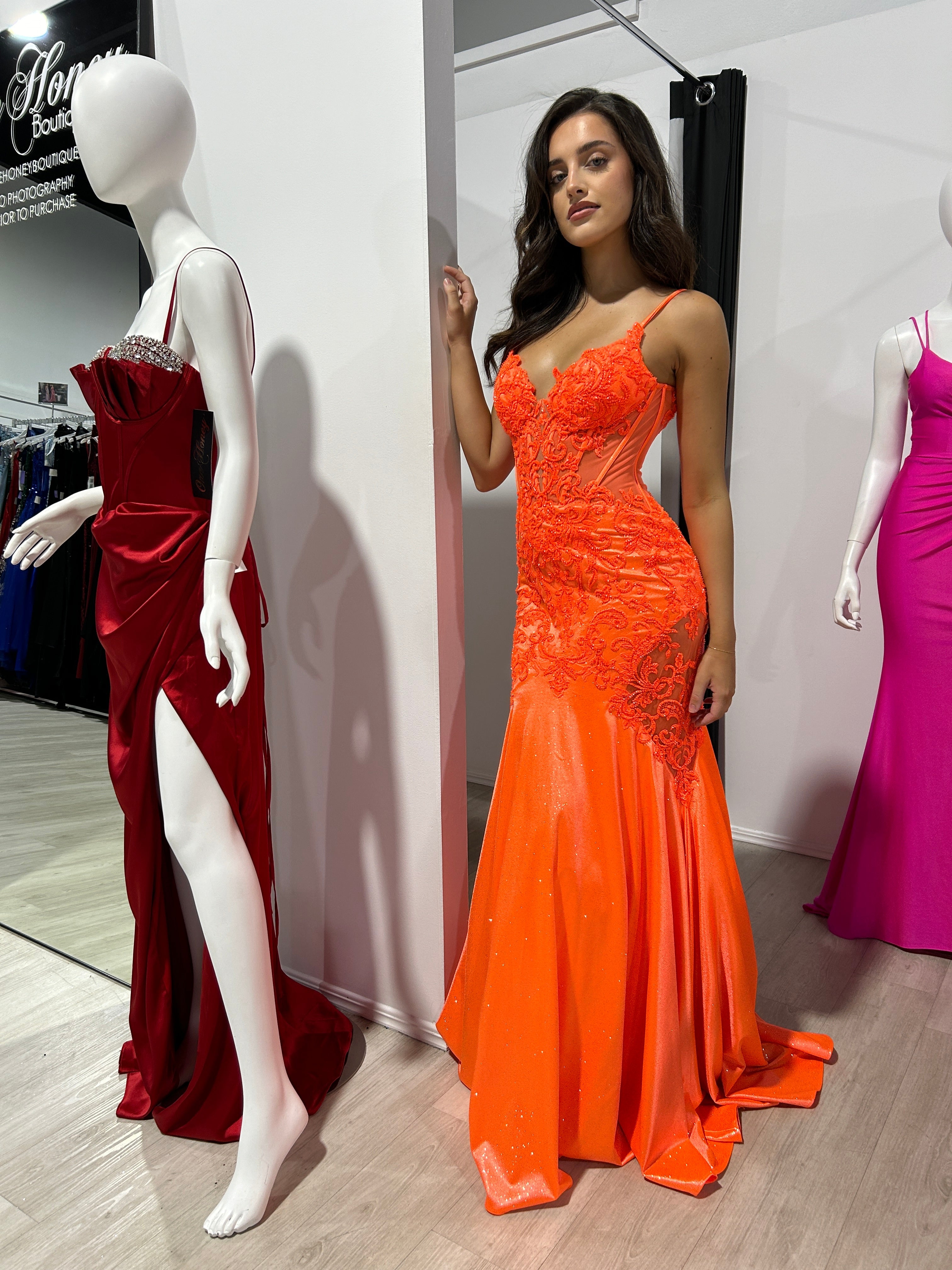 Model wearing an orange lace and satin fitted evening gown, with intricate floral appliqués, standing beside a red dress on a mannequin.