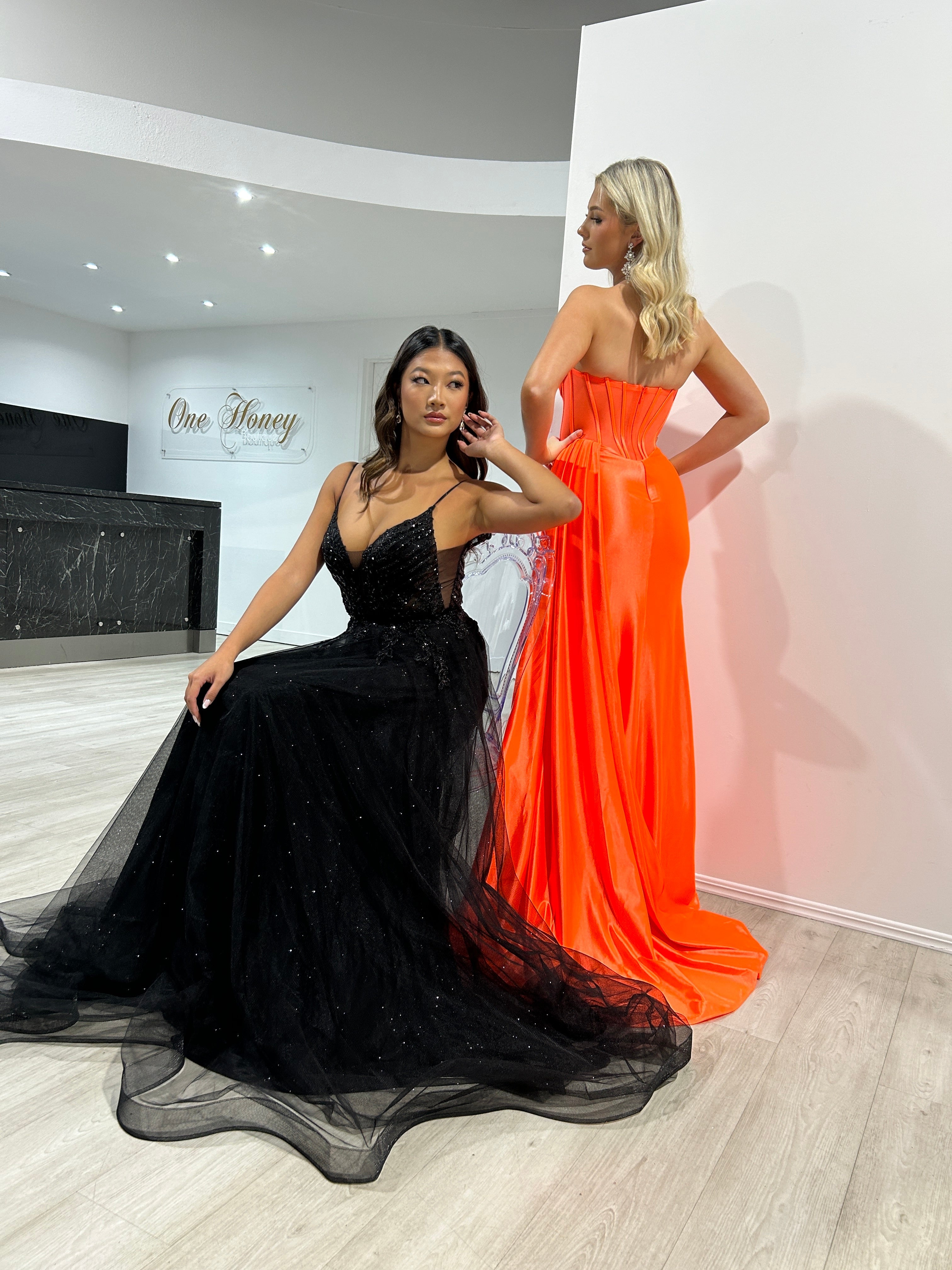 Model in black sequin evening gown sits, while another stands in orange satin strapless dress. Elegant formal wear display.