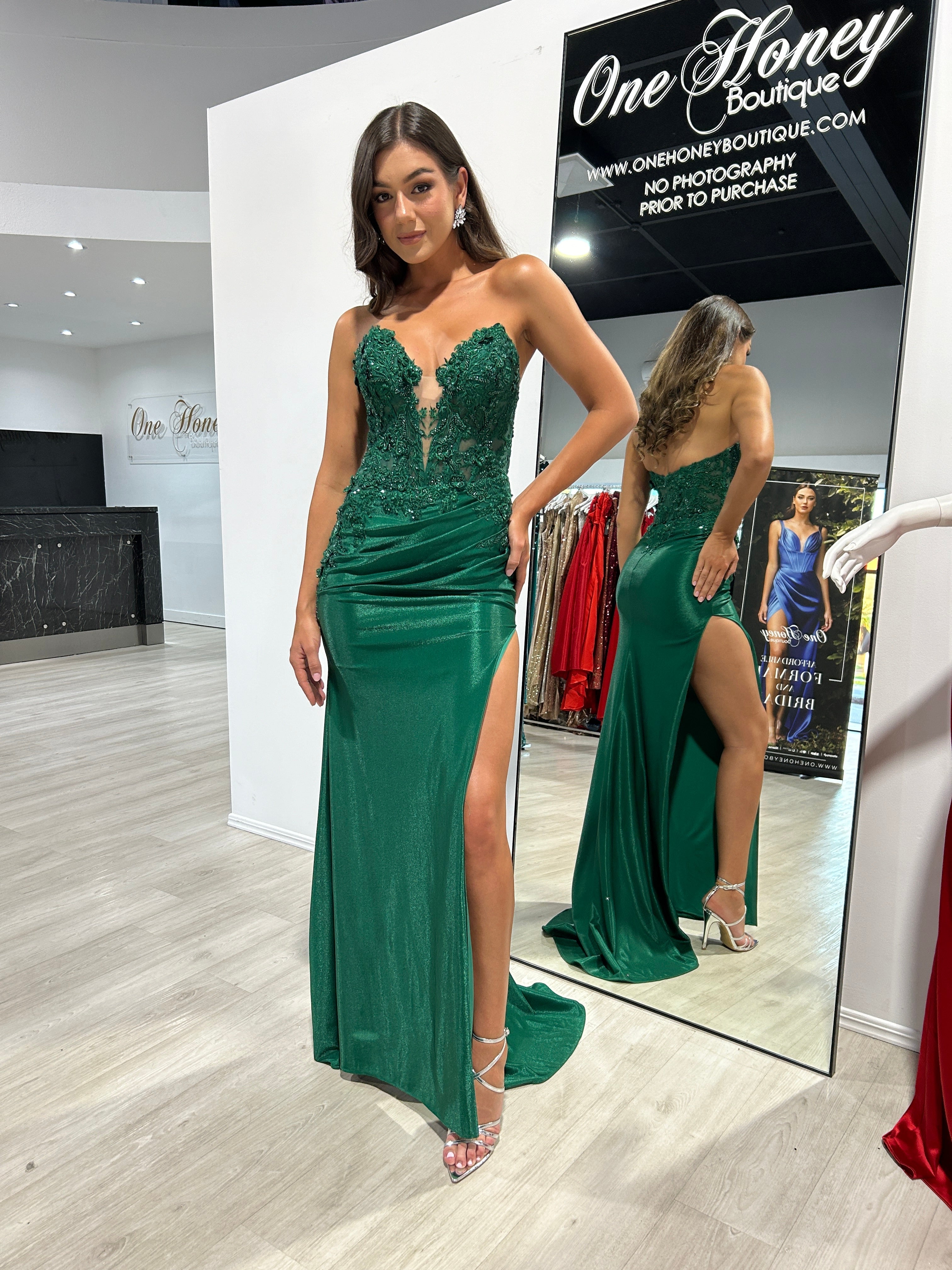 Woman wearing a green strapless gown with lace bodice and high slit, standing in a boutique fitting room. Elegant evening dress.