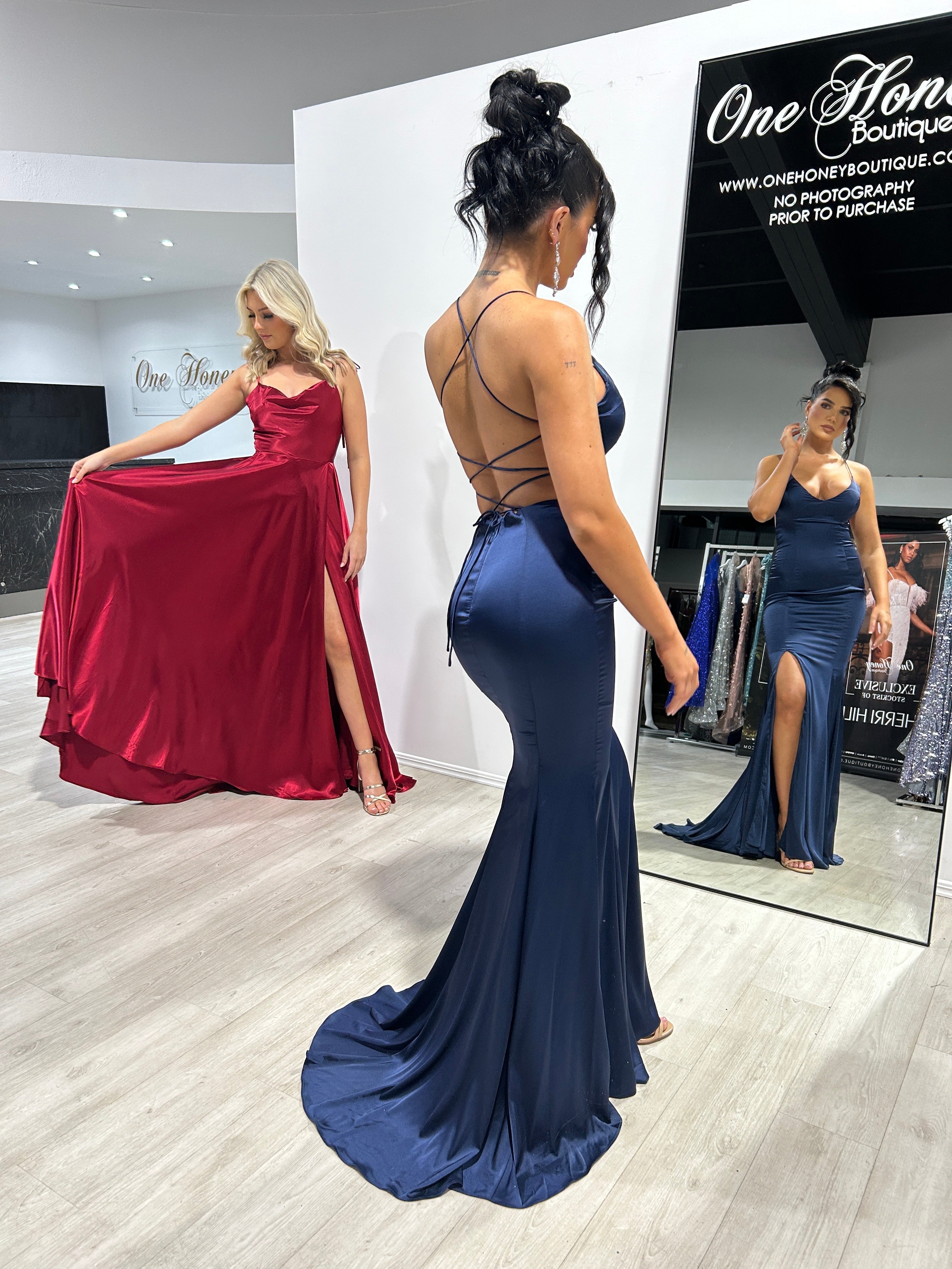 Two women in formal gowns; one wears a red dress with thigh-high slit, the other a navy gown with lace-up back, in a boutique.