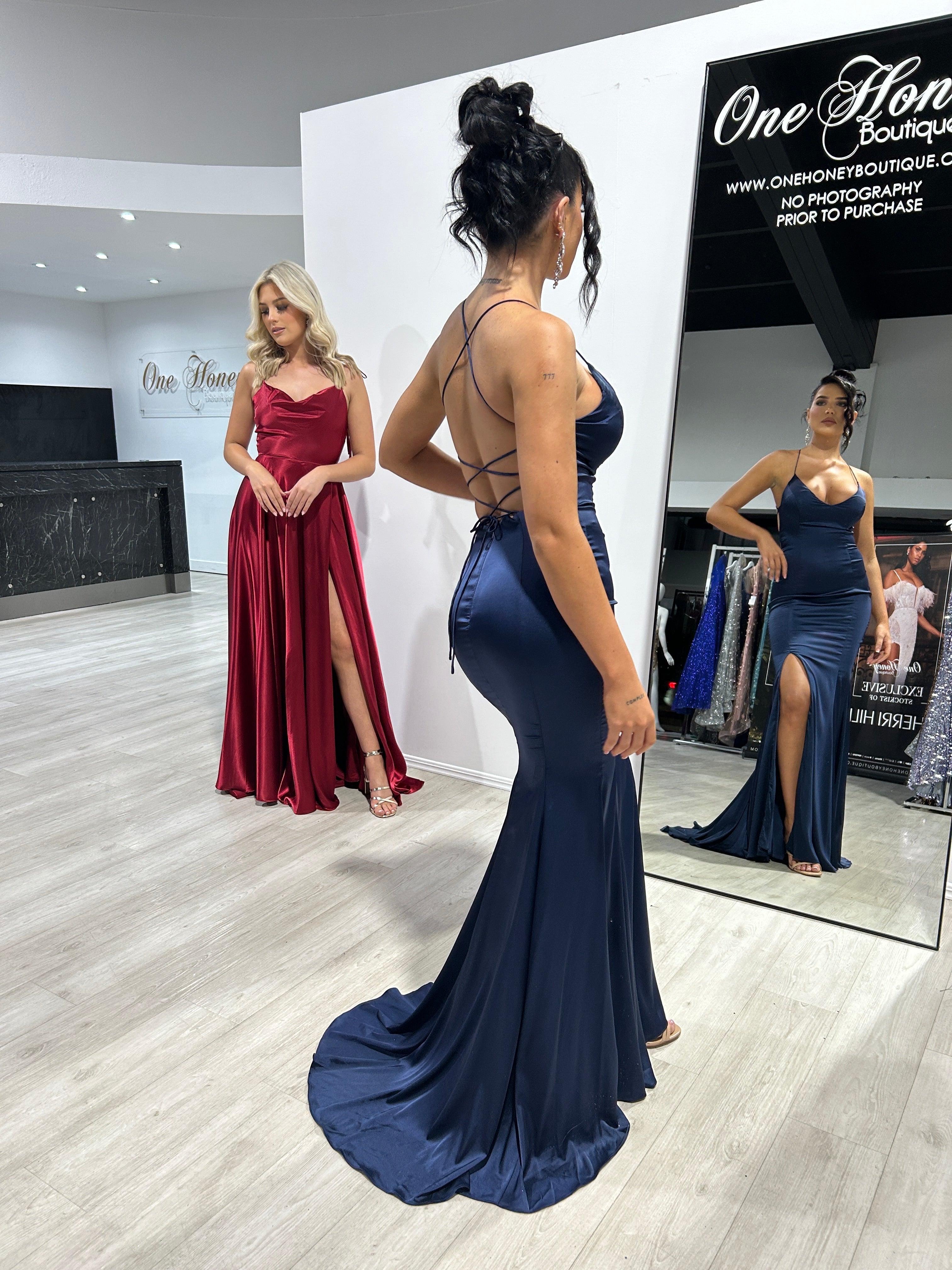 A navy blue mermaid gown with criss-cross back, paired with a red satin dress featuring a thigh-high slit. Evening wear in boutique setting.