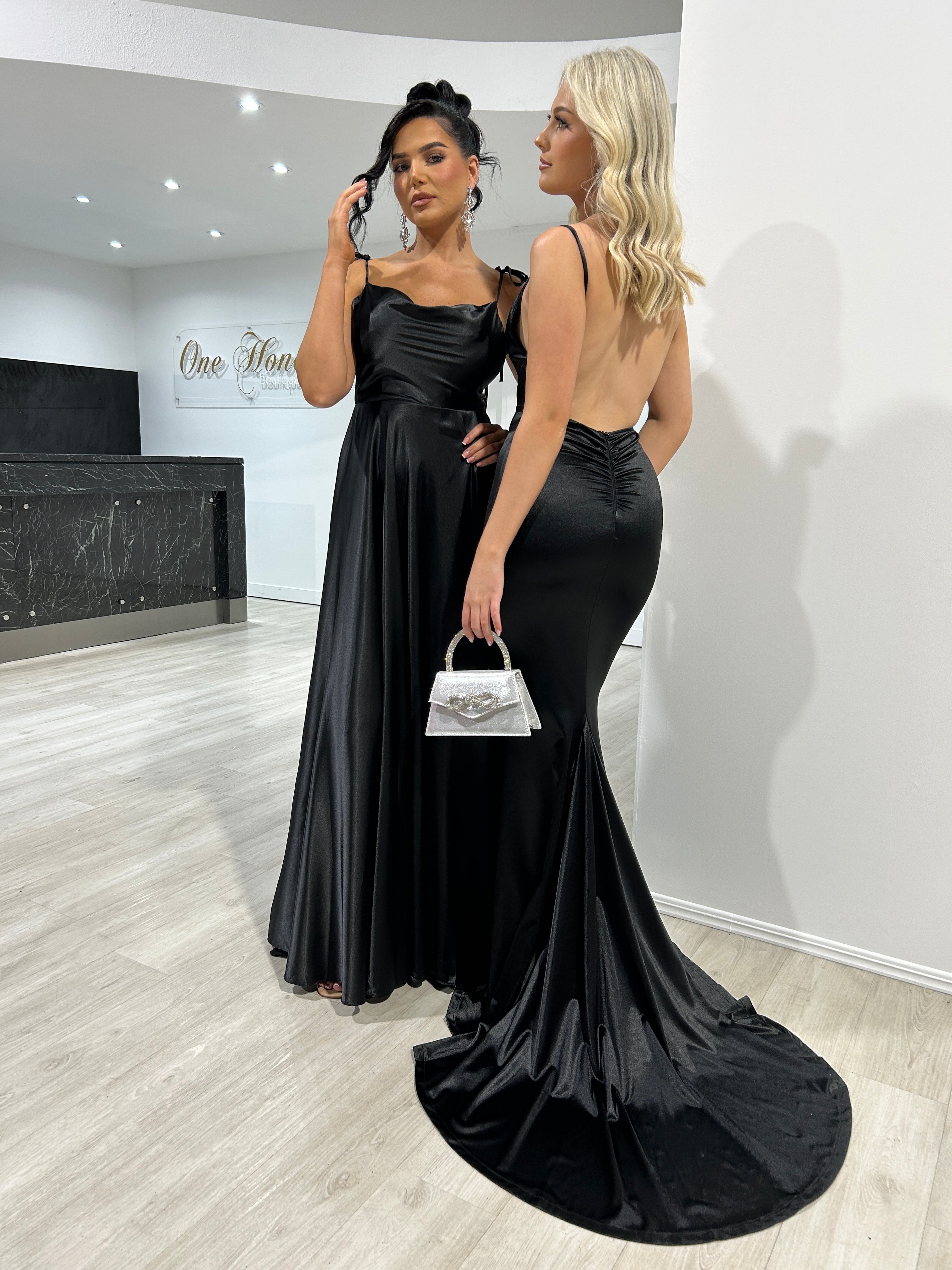 Two women in elegant black Honey Couture KYLIE evening gowns, one with a low back mermaid design, in a chic showroom – Australian Designer Prom Dresses.