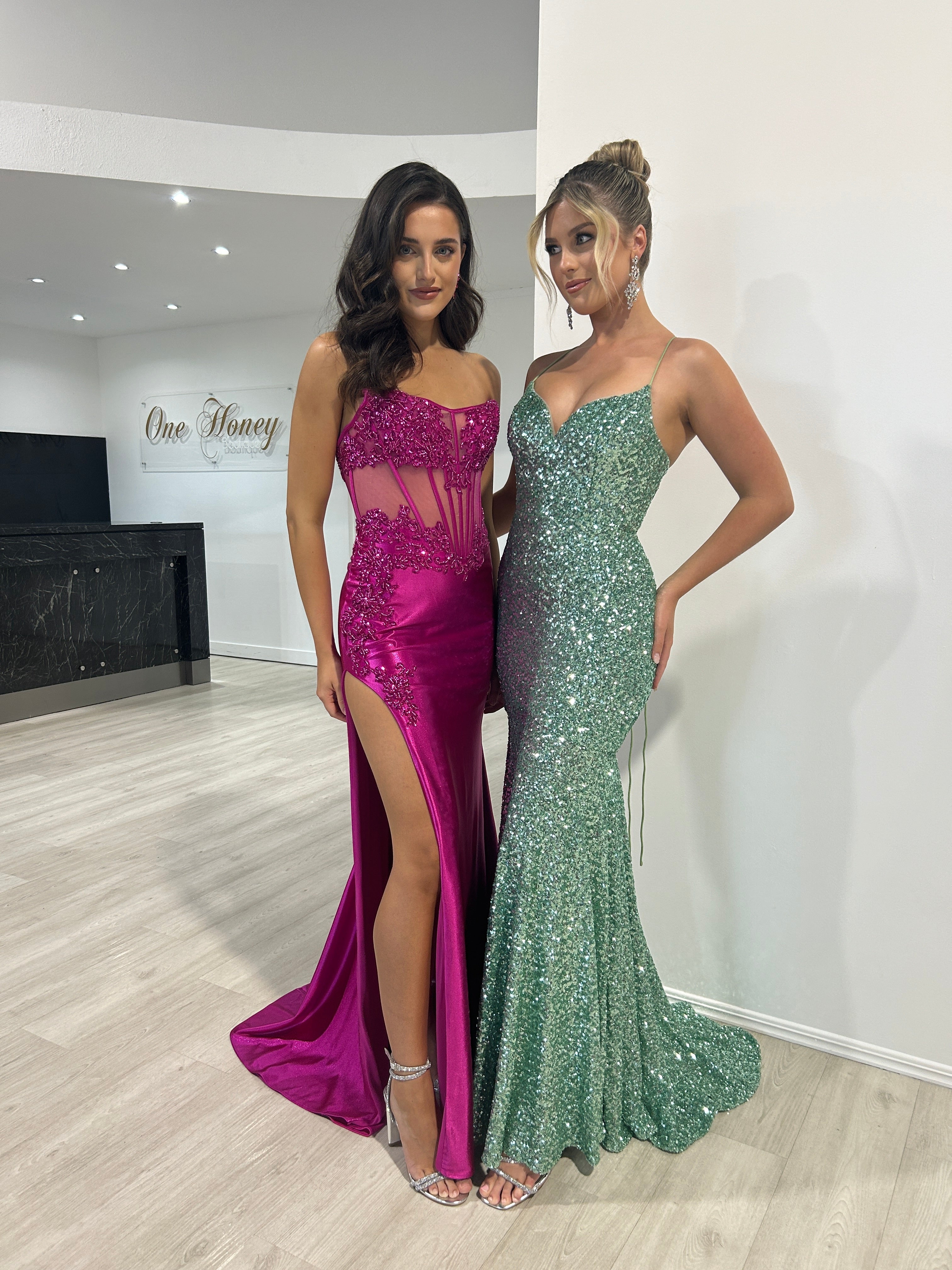 Two women in elegant evening gowns, one in a fuchsia sheer corset dress with thigh-high slit, and the other in a shimmering green gown. Brisbane to Gold Coast.