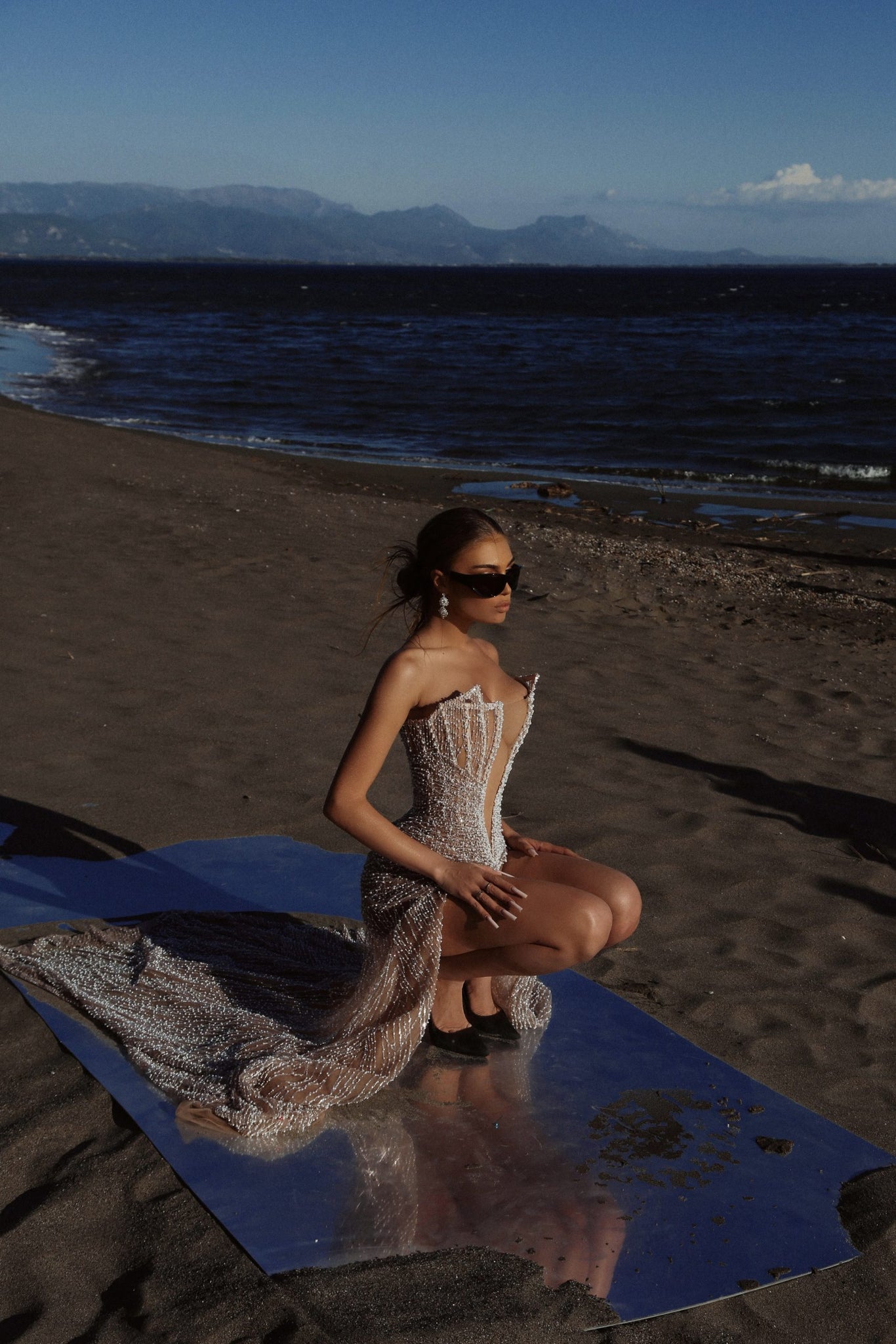 Woman wearing a strapless beaded gown with a mermaid tail on a reflective surface at the beach.
