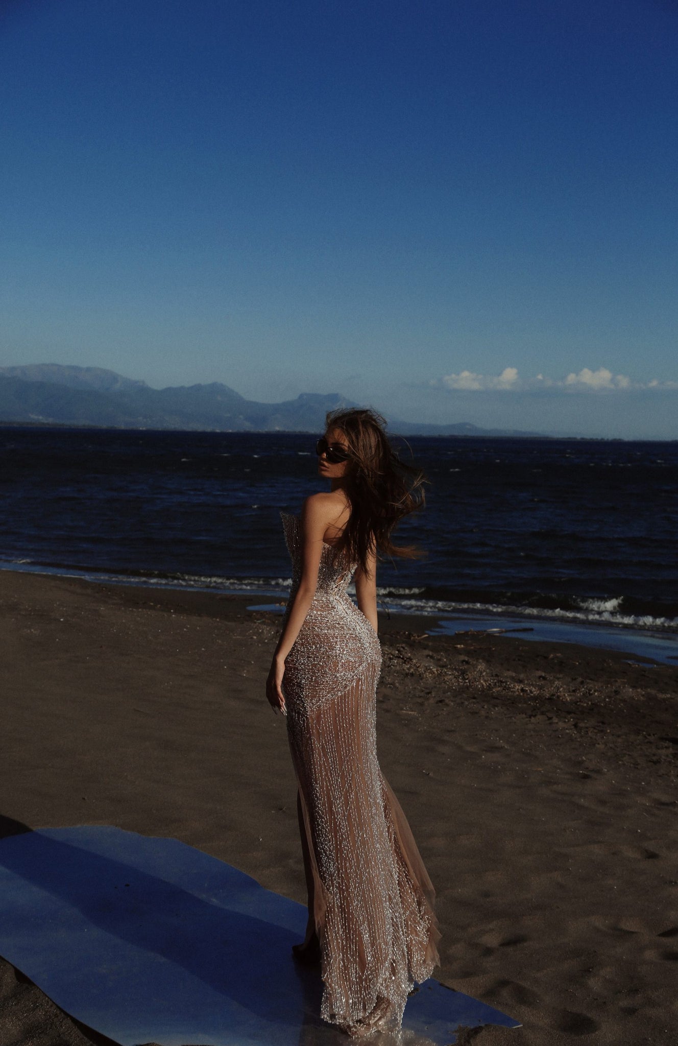 Woman in a nude beaded mermaid dress on a beach with waves and mountains in the background, embracing elegance and sophistication.