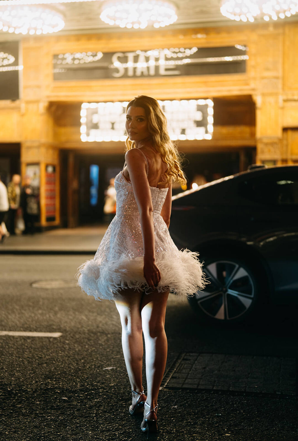 Woman wearing a white sequin and feather mini corset dress, standing on a street at night, perfect for a hens party or bridal event.