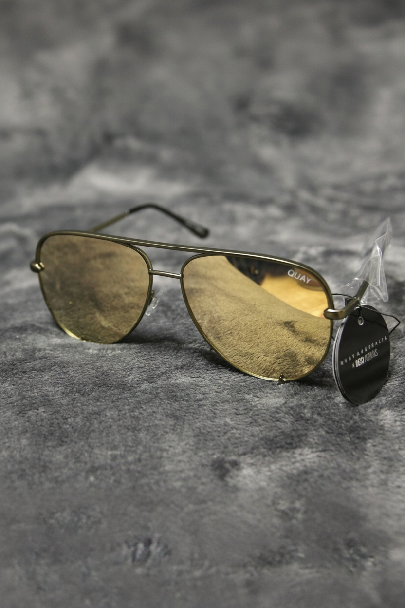 A pair of aviator-style sunglasses with gold-toned metal frames and tinted lenses, resting on a textured gray surface. A black hangtag is attached to the right side, and the brand name 'QUAY' is visible on the upper frame.