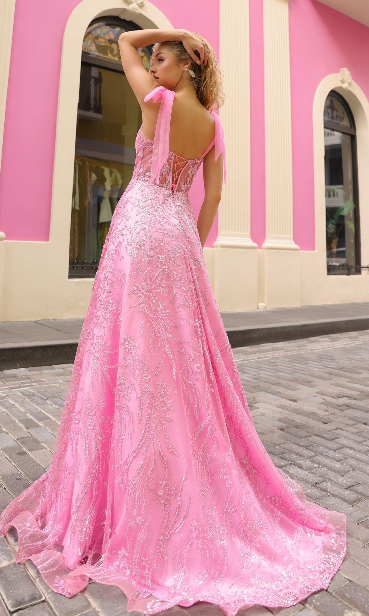 Woman wearing a pink sequin ball gown with tulle shoulder bows, featuring a sheer corset-style bodice, on a cobblestone street.