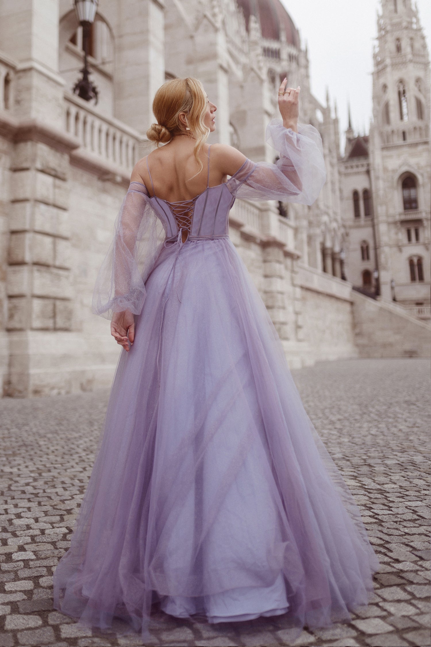 Woman in lavender long sleeve ball gown with sheer sleeves walking near historic building, showcasing elegant formal wear style.