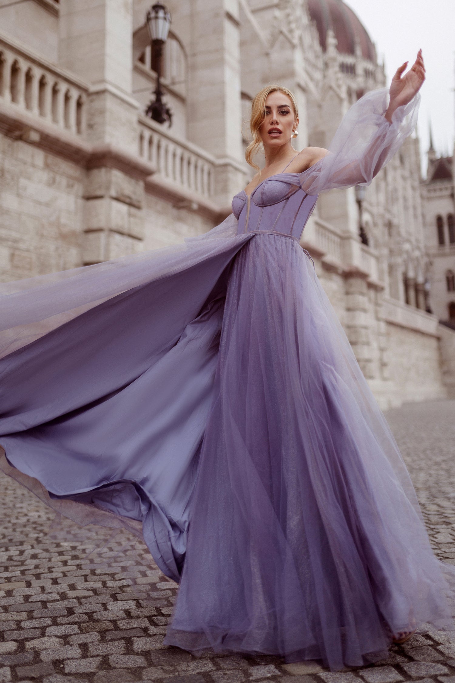 Enchanting purple tulle ball gown with off-shoulder sleeves, flowing gracefully on cobblestone street. Perfect for formal events.