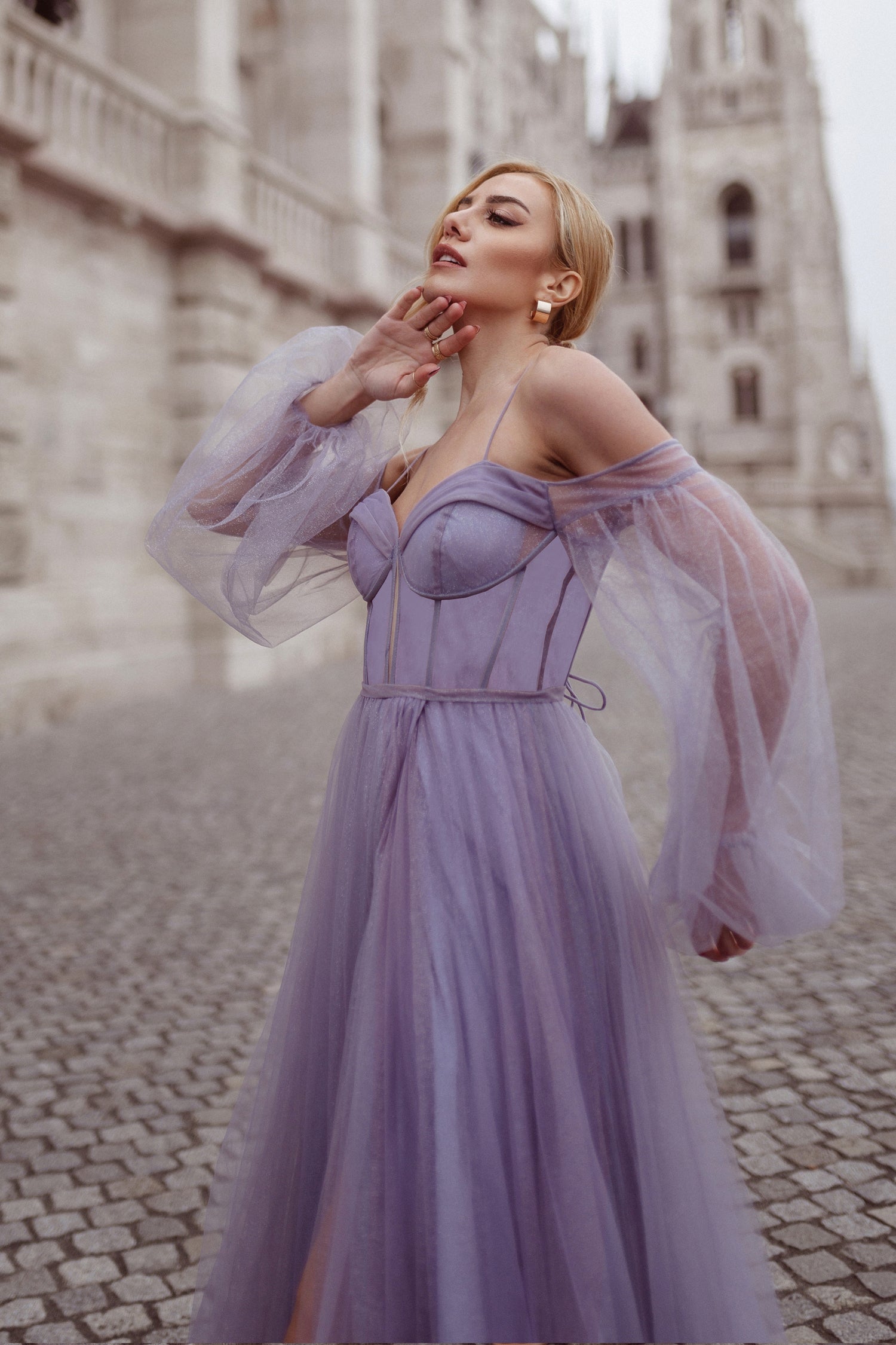 Elegant woman in a purple tulle ball gown with sheer puffy sleeves, posing on cobblestone street beside a historic building.