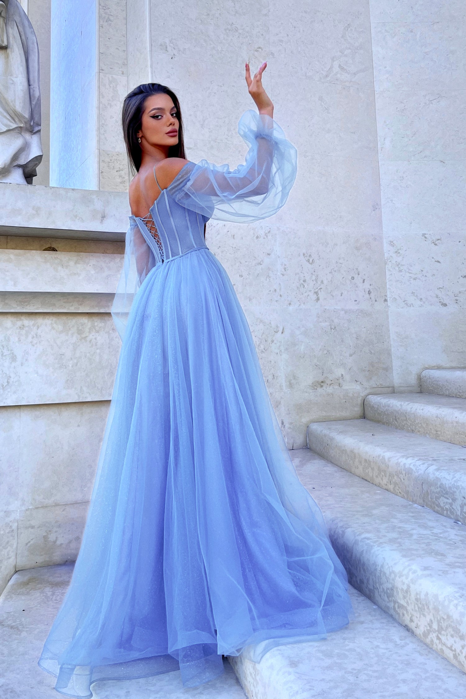 Elegant blue long sleeve formal dress with sheer sleeves and fitted bodice, worn by woman on stone stairs.