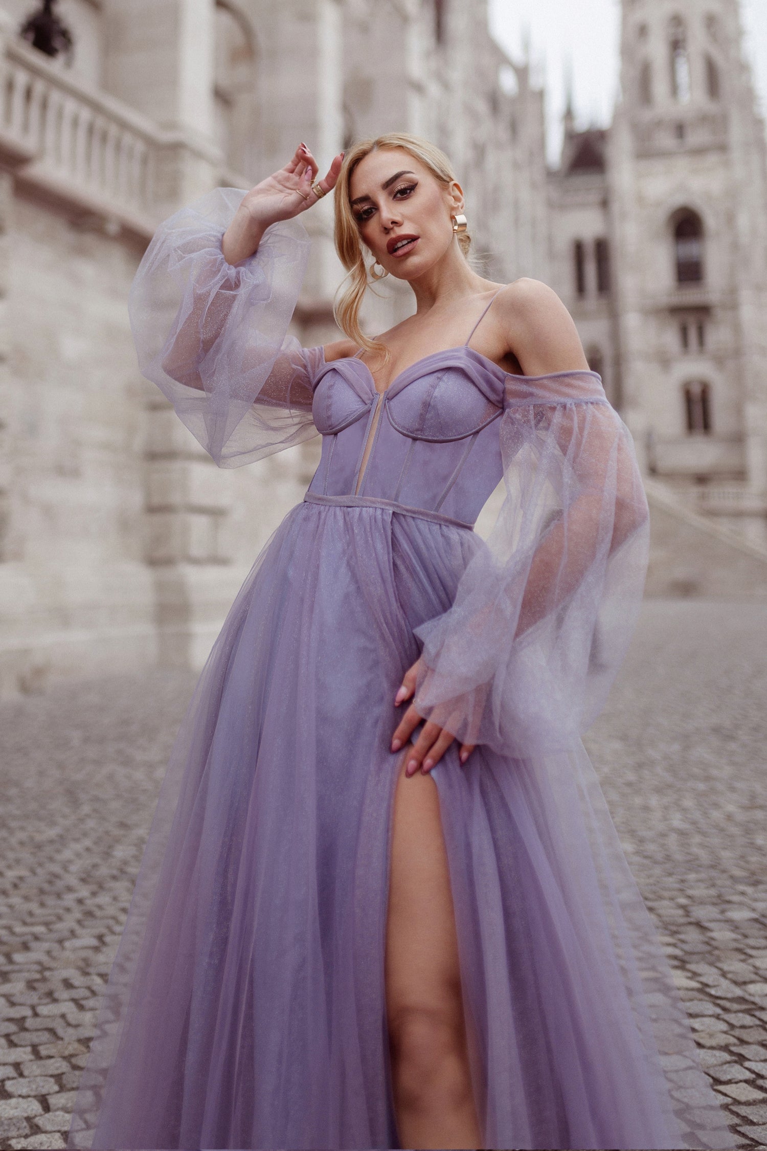 Lavender off-shoulder ball gown with sheer puff sleeves and leg slit, perfect as an evening dress, set against a historic building backdrop.