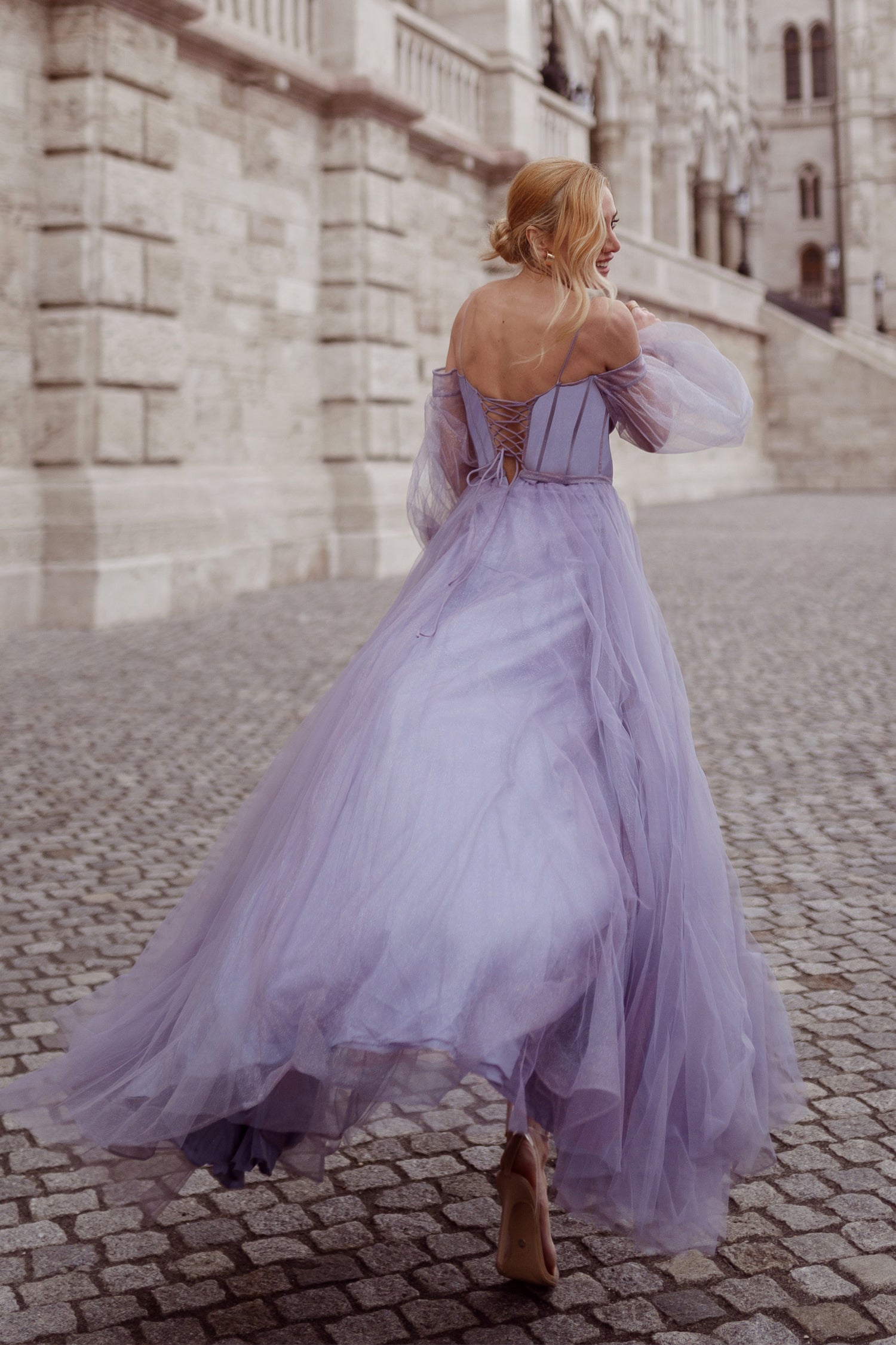 Woman in a flowing lavender ball gown walking on cobblestone street near a historic building.