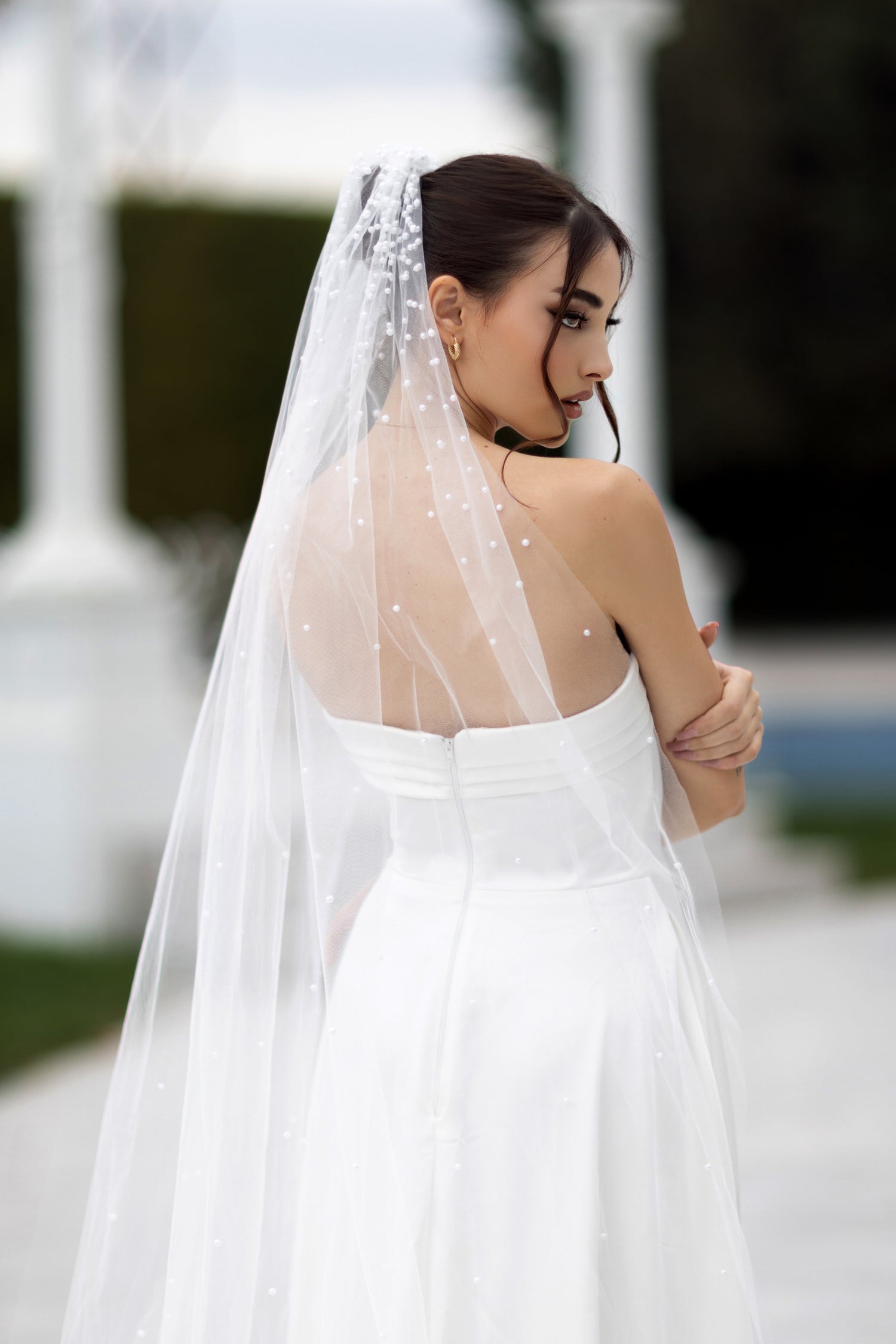 Bride in strapless wedding gown with long white veil, outdoor setting.