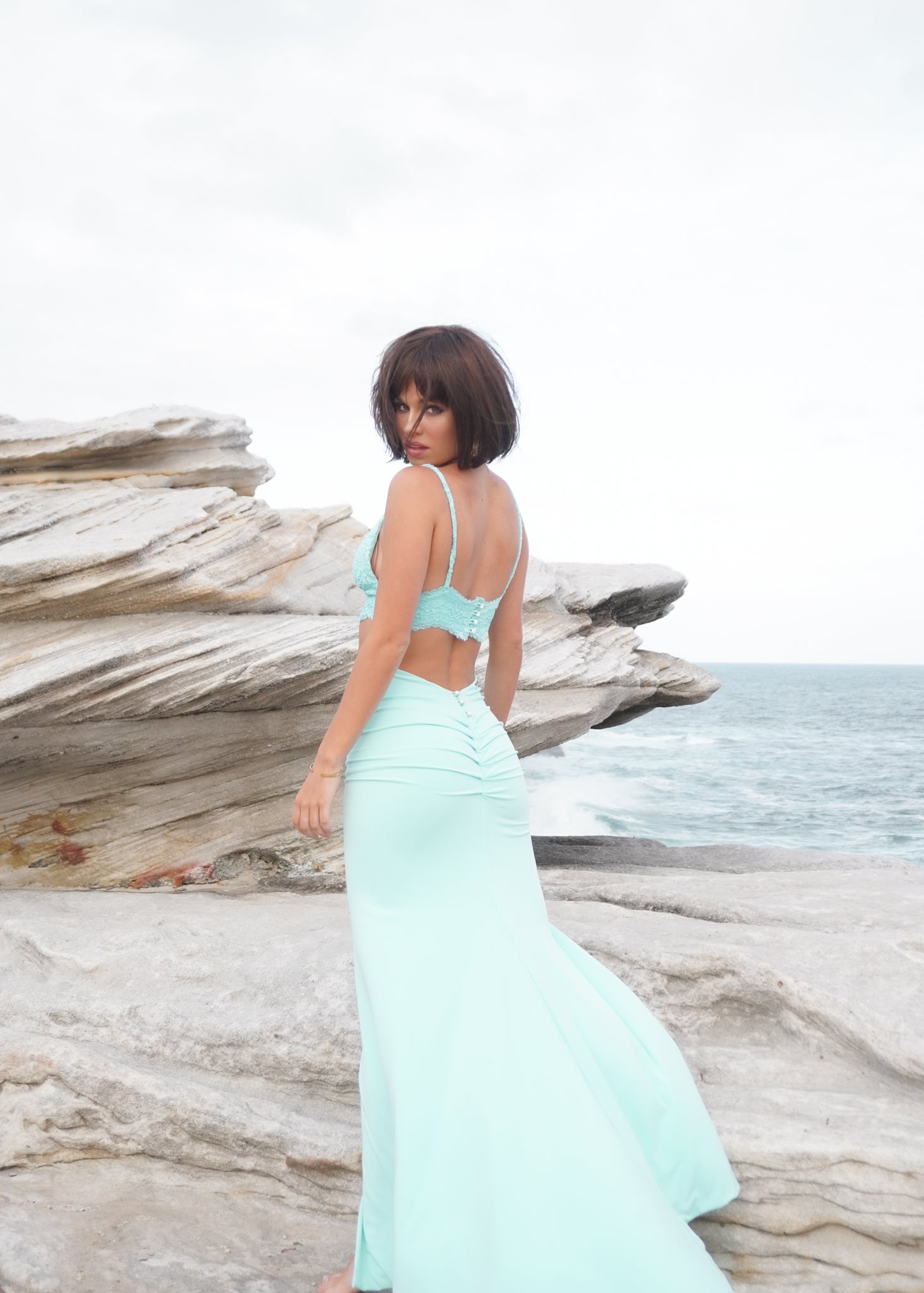 Elegant woman in aqua evening dress on a coastal rock, showcasing back view of a fitting gown with natural scenery.