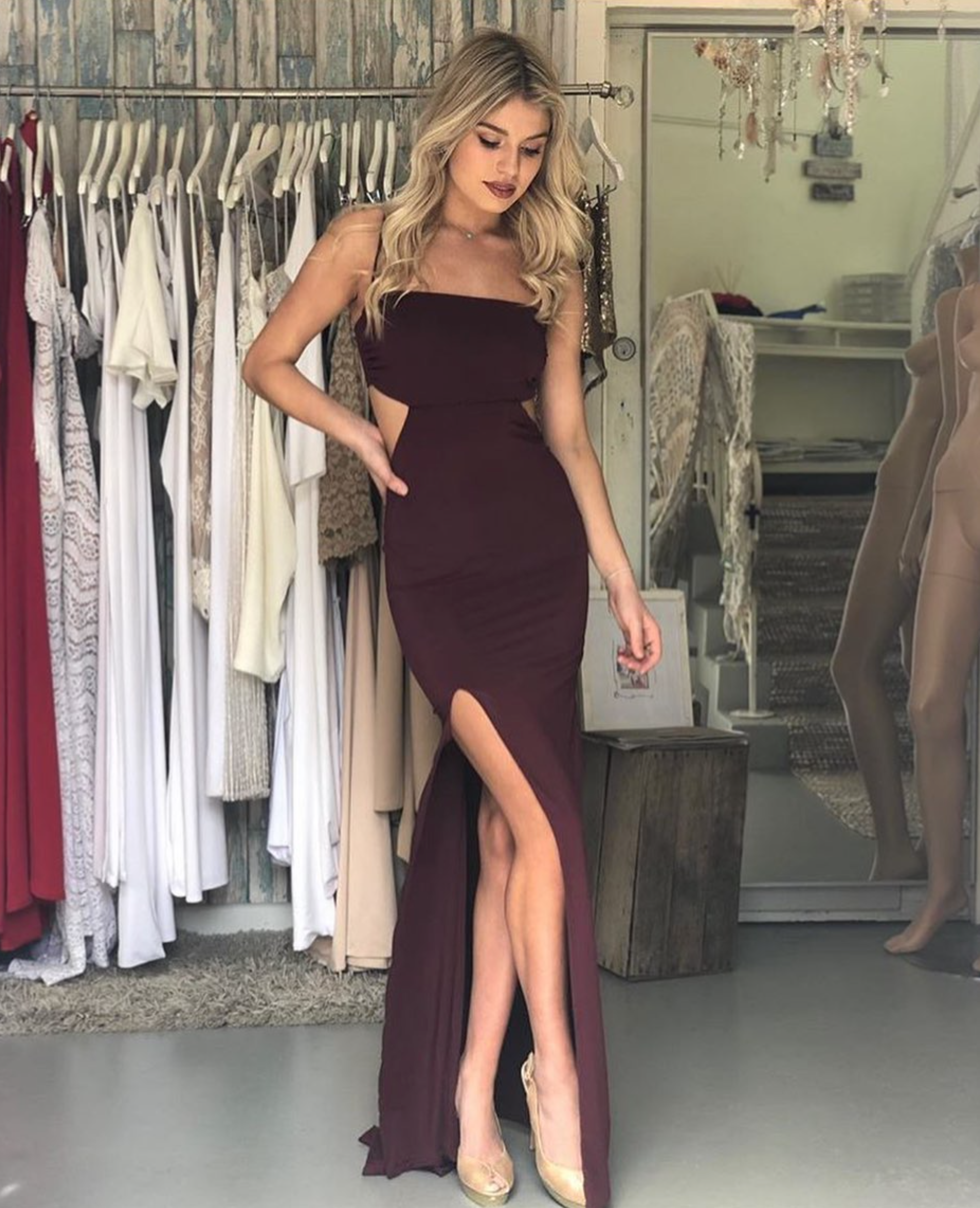 Woman wearing burgundy strapless formal gown with leg split, surrounded by dresses in boutique.