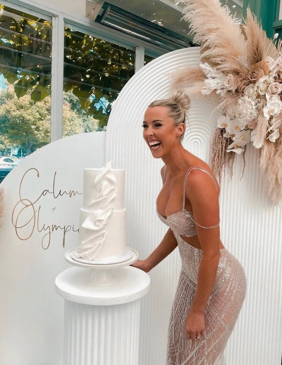 Woman in a sparkling formal dress smiling at a white tiered cake on a pedestal with floral decor in the background.