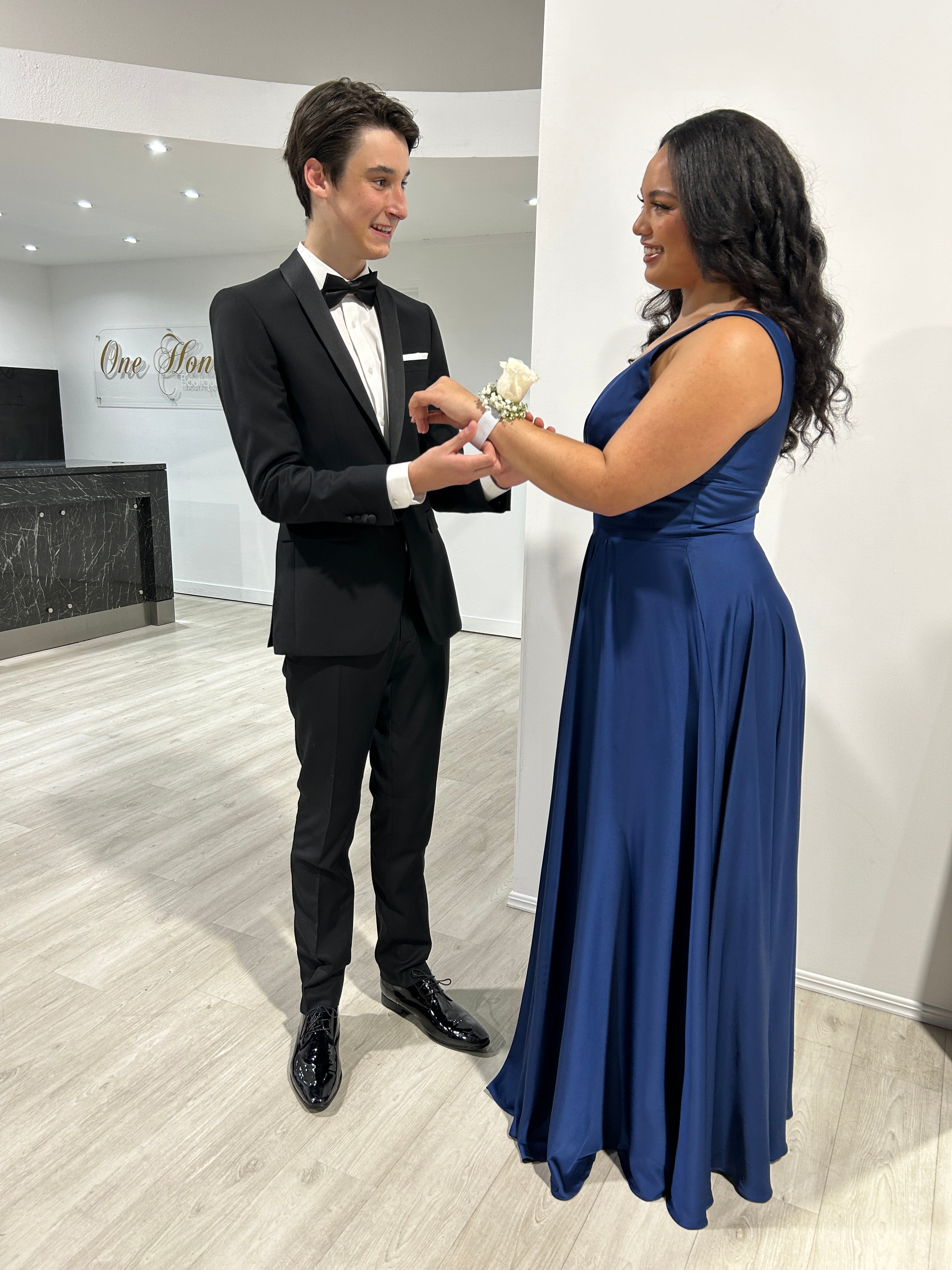 Young couple at a formal event, man in a black tuxedo, woman in a blue evening gown receiving a corsage, standing in a modern venue.