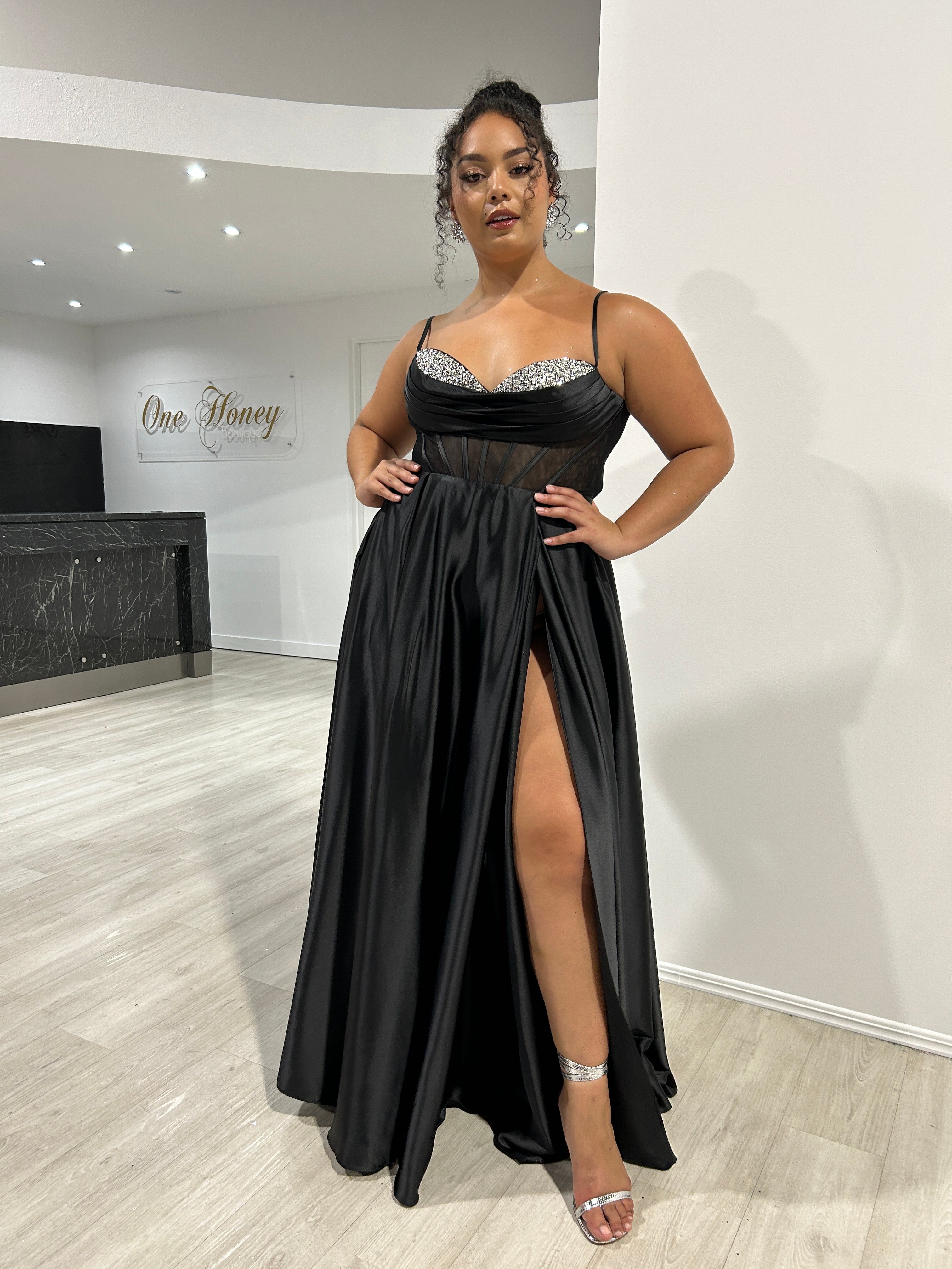 Woman wearing black satin formal gown with diamanté detailed bust, featuring a front slit, in a stylish boutique setting.