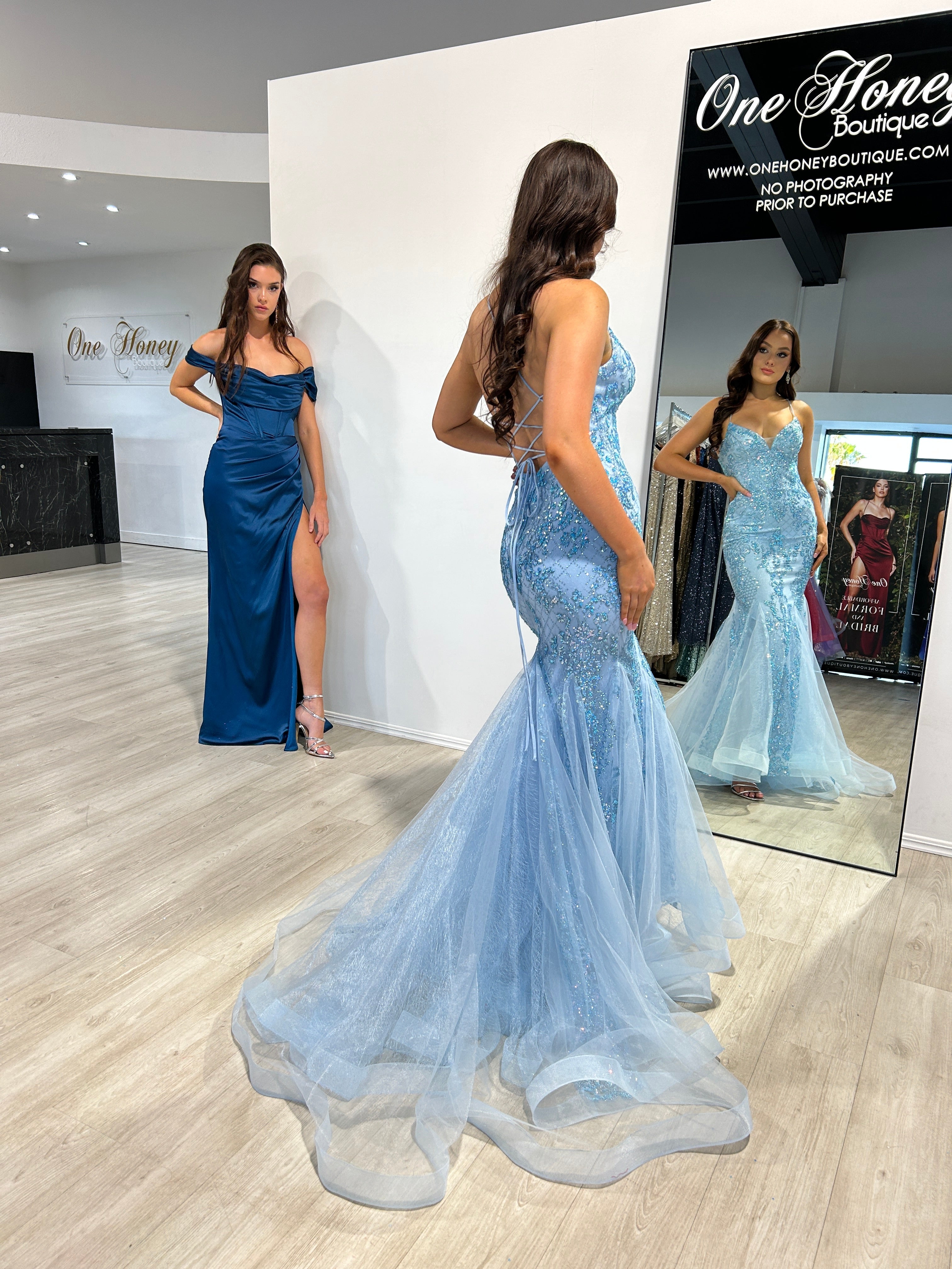 Woman in blue strapless mermaid prom gown with lace bodice looks in mirror; another woman in an off-shoulder navy gown stands nearby. Brisbane to Gold Coast.