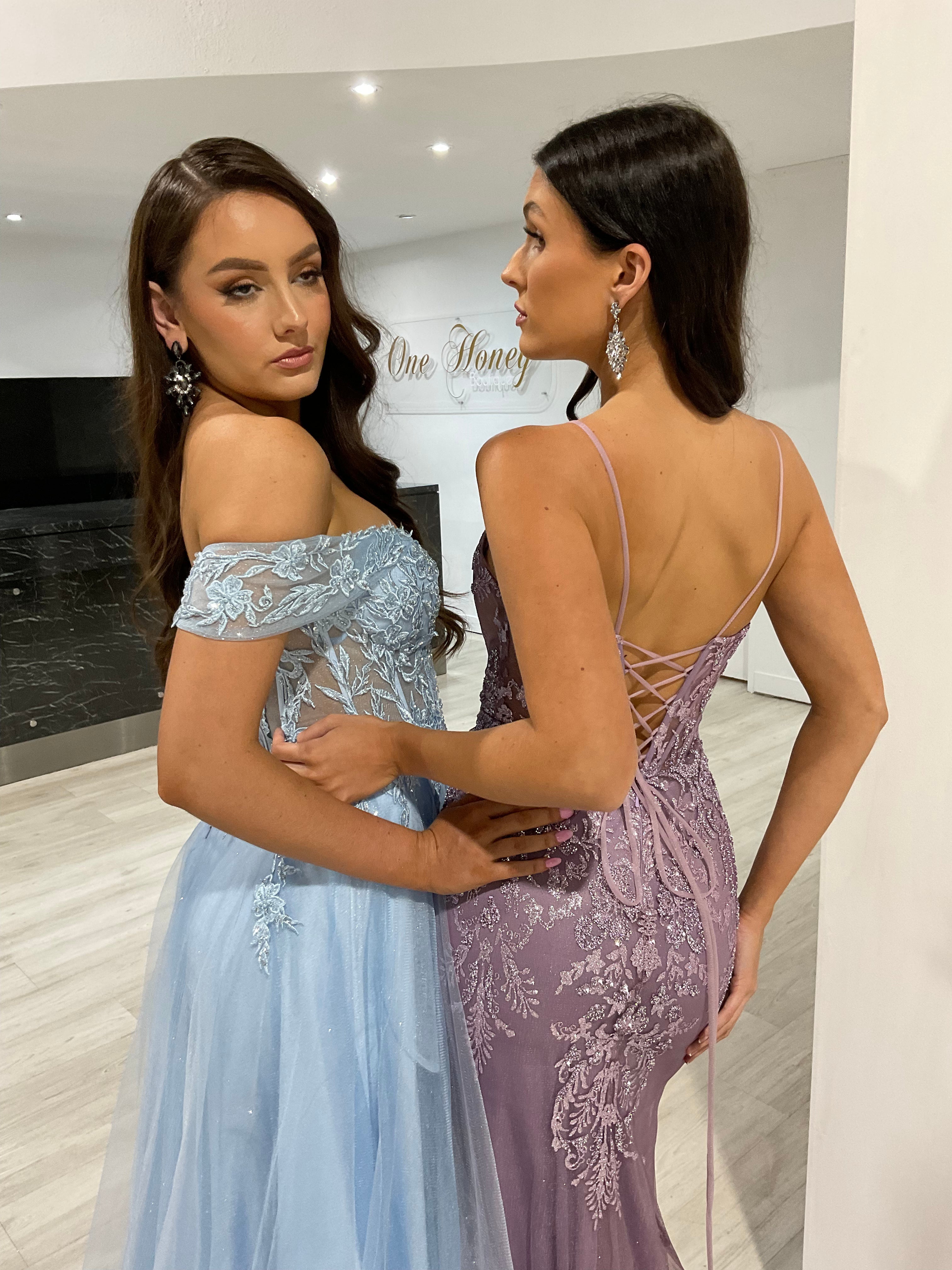 Two women in elegant lace formal gowns with floral embroidery and corset backs, standing in a boutique showroom. Brisbane to Gold Coast.