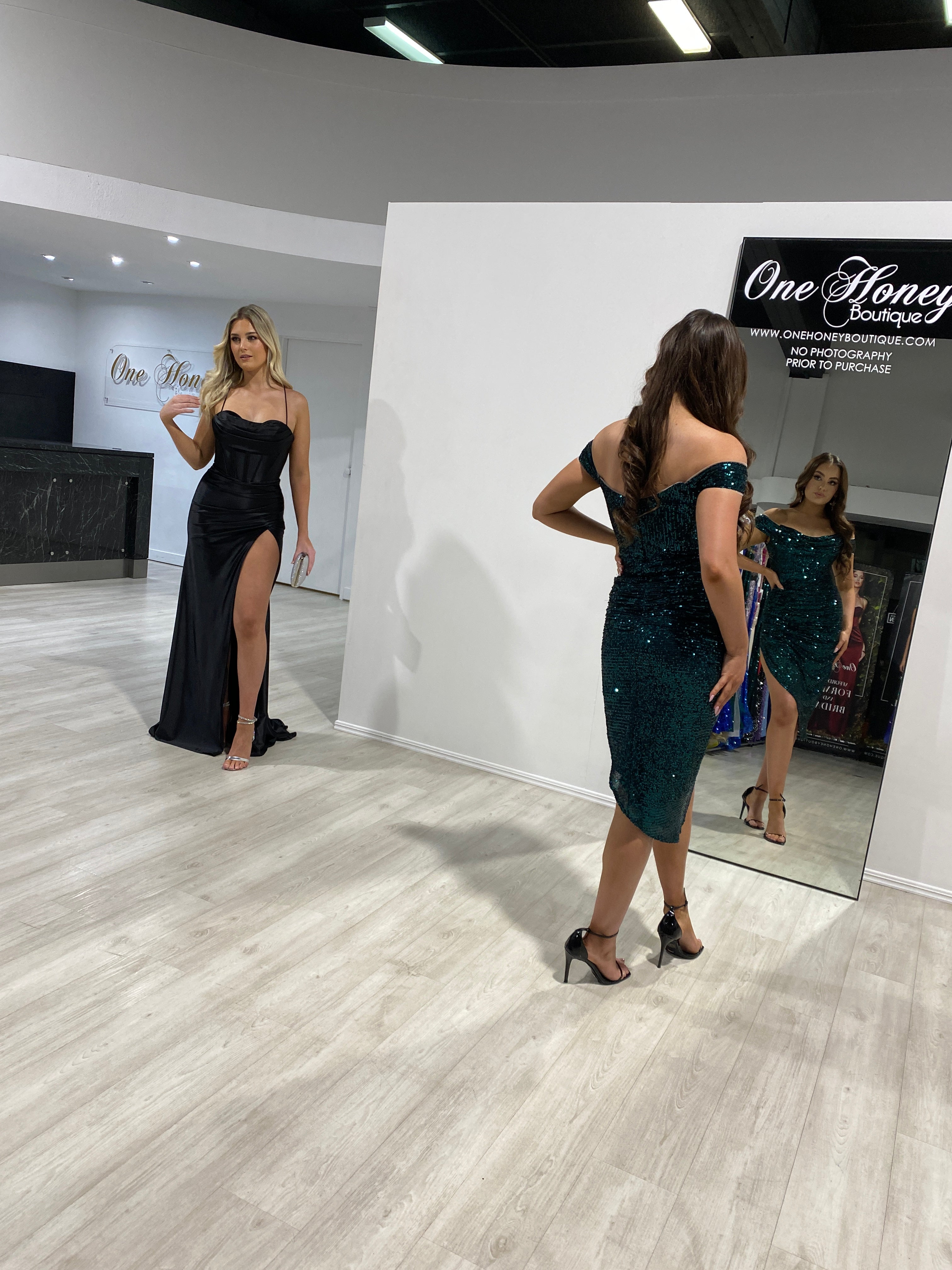 Two women in formal dresses at One Honey Boutique, one in a black slit gown and another in a green sequin dress, in a stylish showroom.