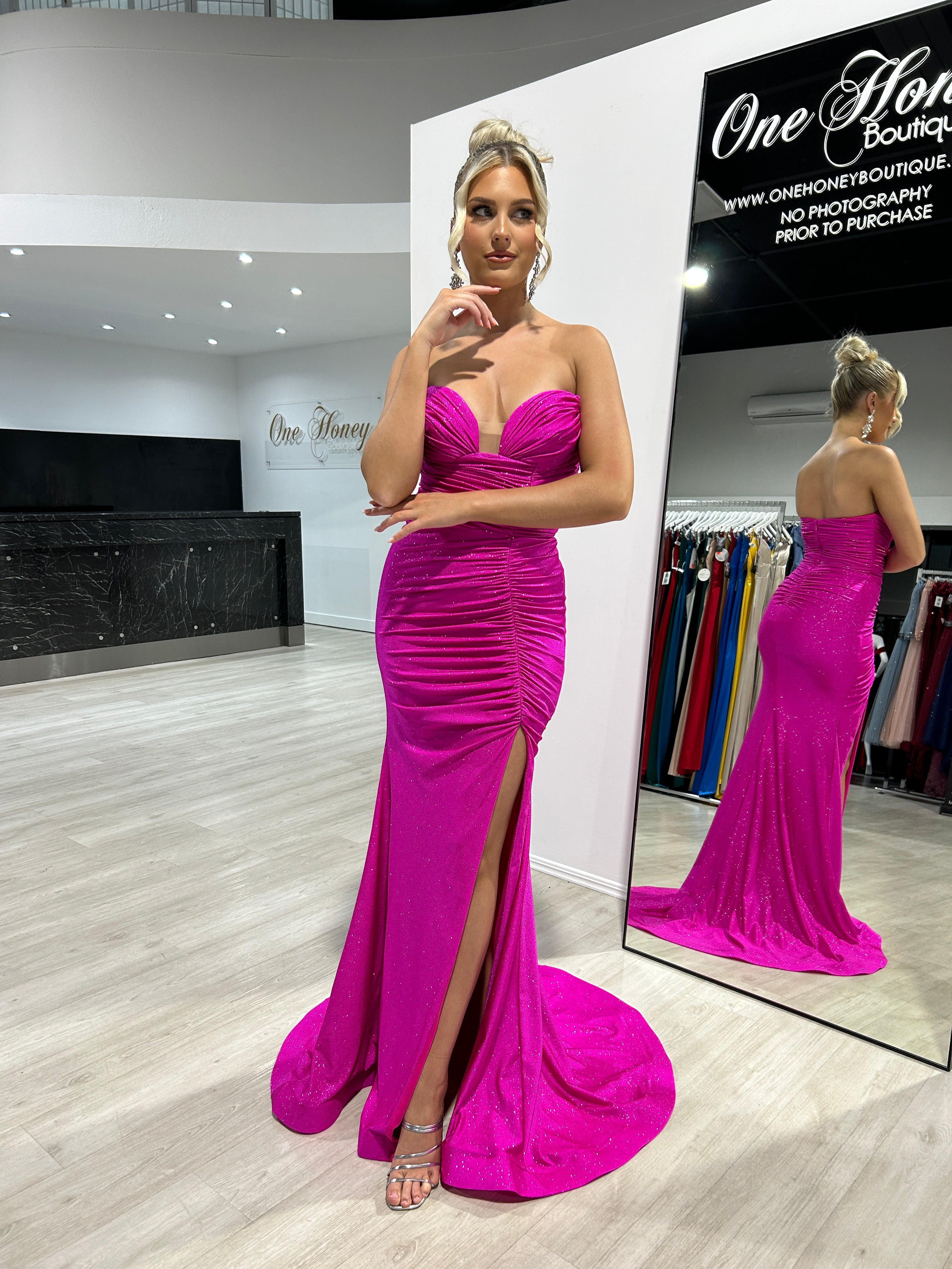 Model wearing a vibrant pink strapless gown with a ruched bodice and side slit, standing in front of a mirror at One Honey Boutique.