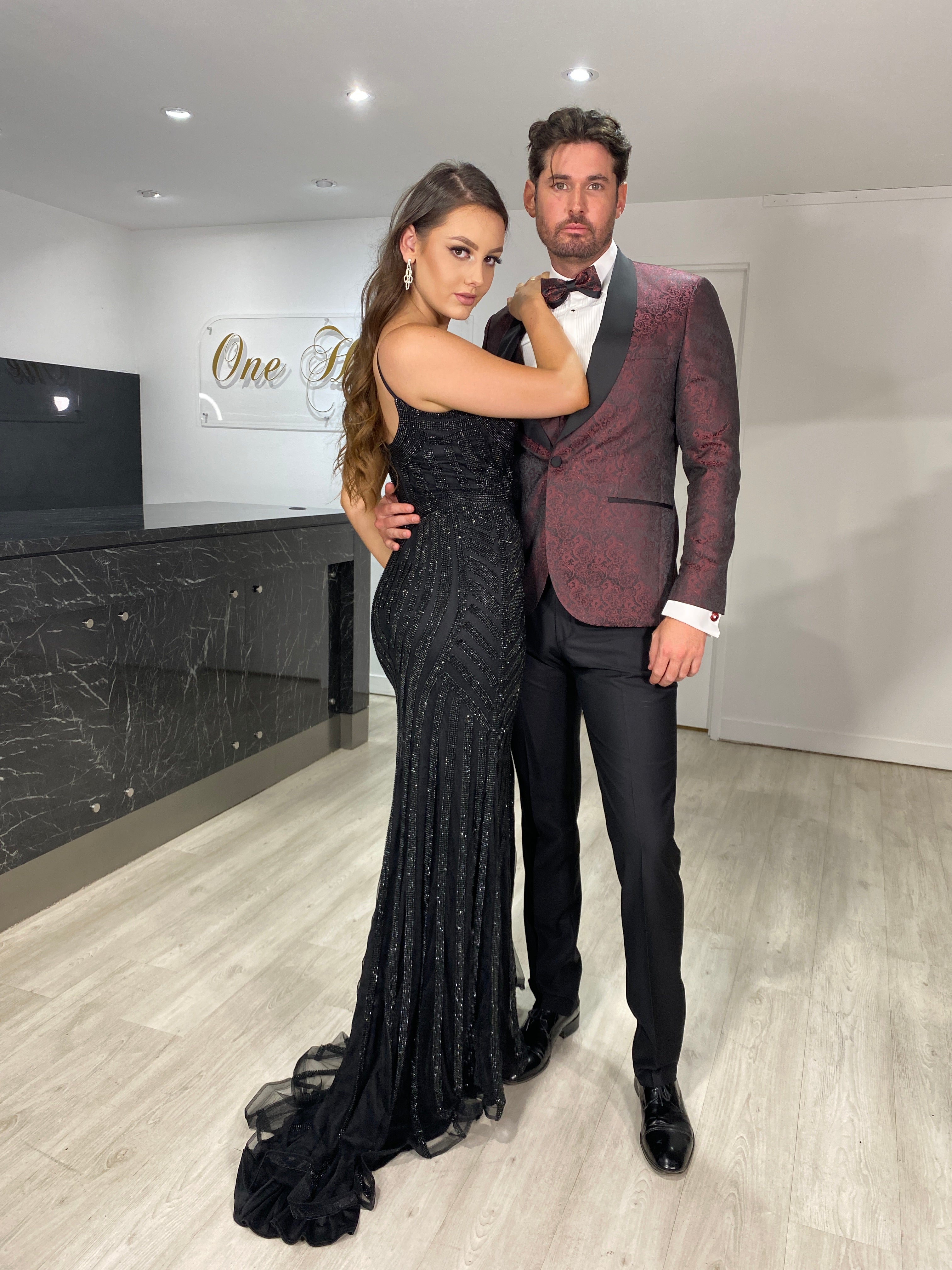 Couple in formal attire; woman in black sequin gown, man in burgundy suit jacket, standing in elegantly designed interior space.