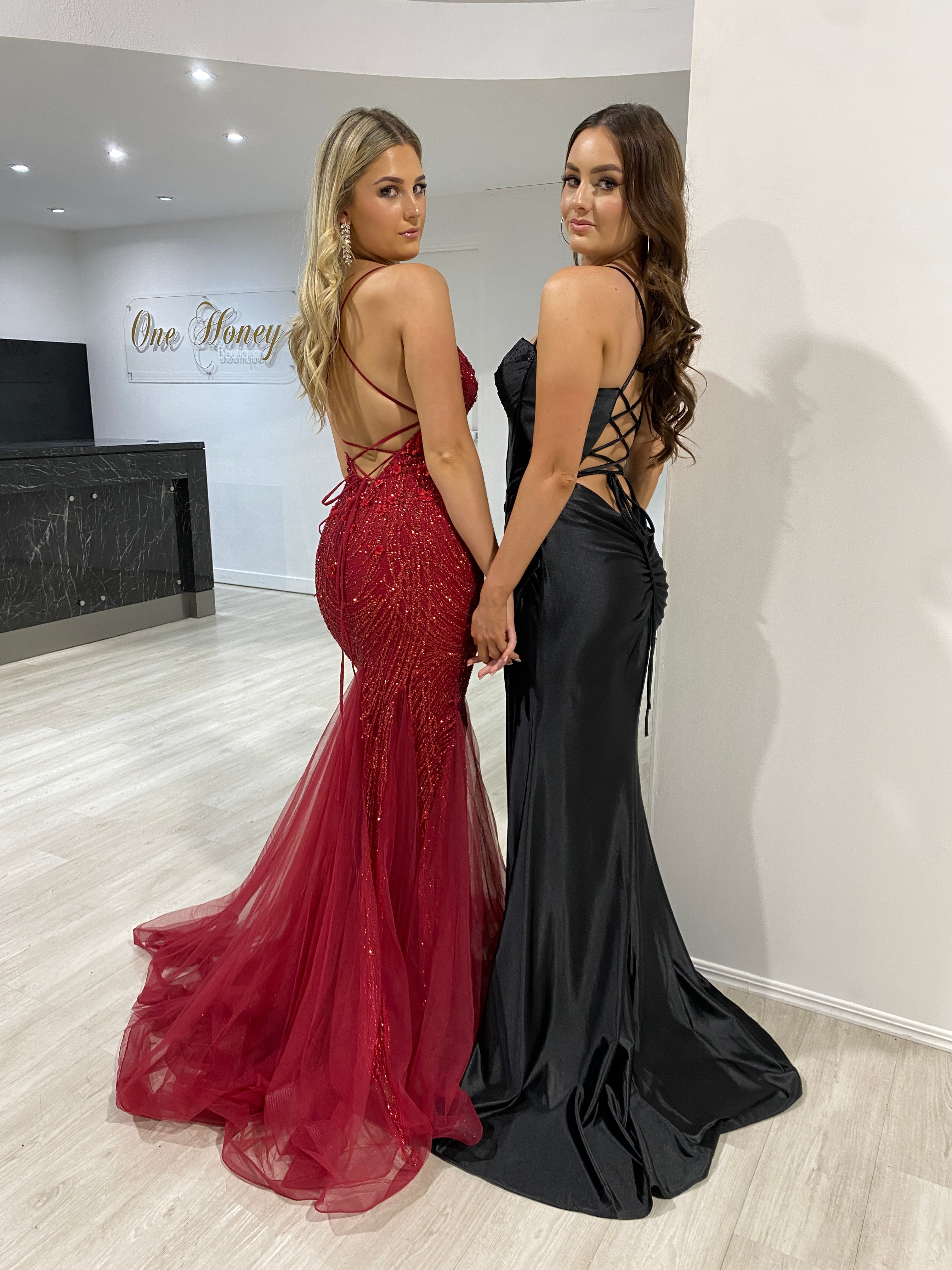 Two women in backless lace-up formal mermaid gowns, one in red glitter and one in black satin, pose elegantly. Brisbane to Gold Coast.