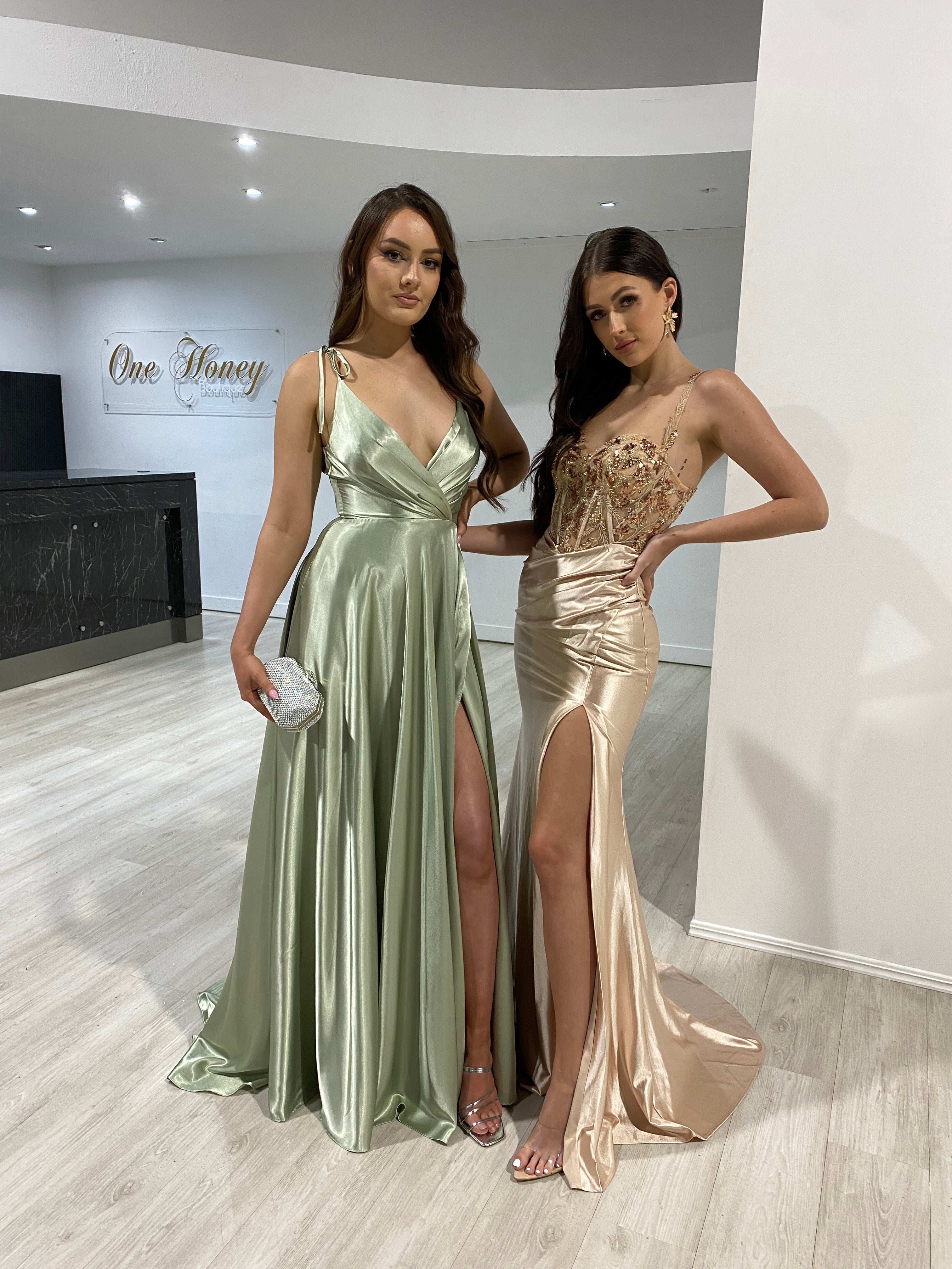Two women wearing Honey Couture silky A Line formal dresses in sage green and champagne, featuring bow tie straps and hidden leg splits.
