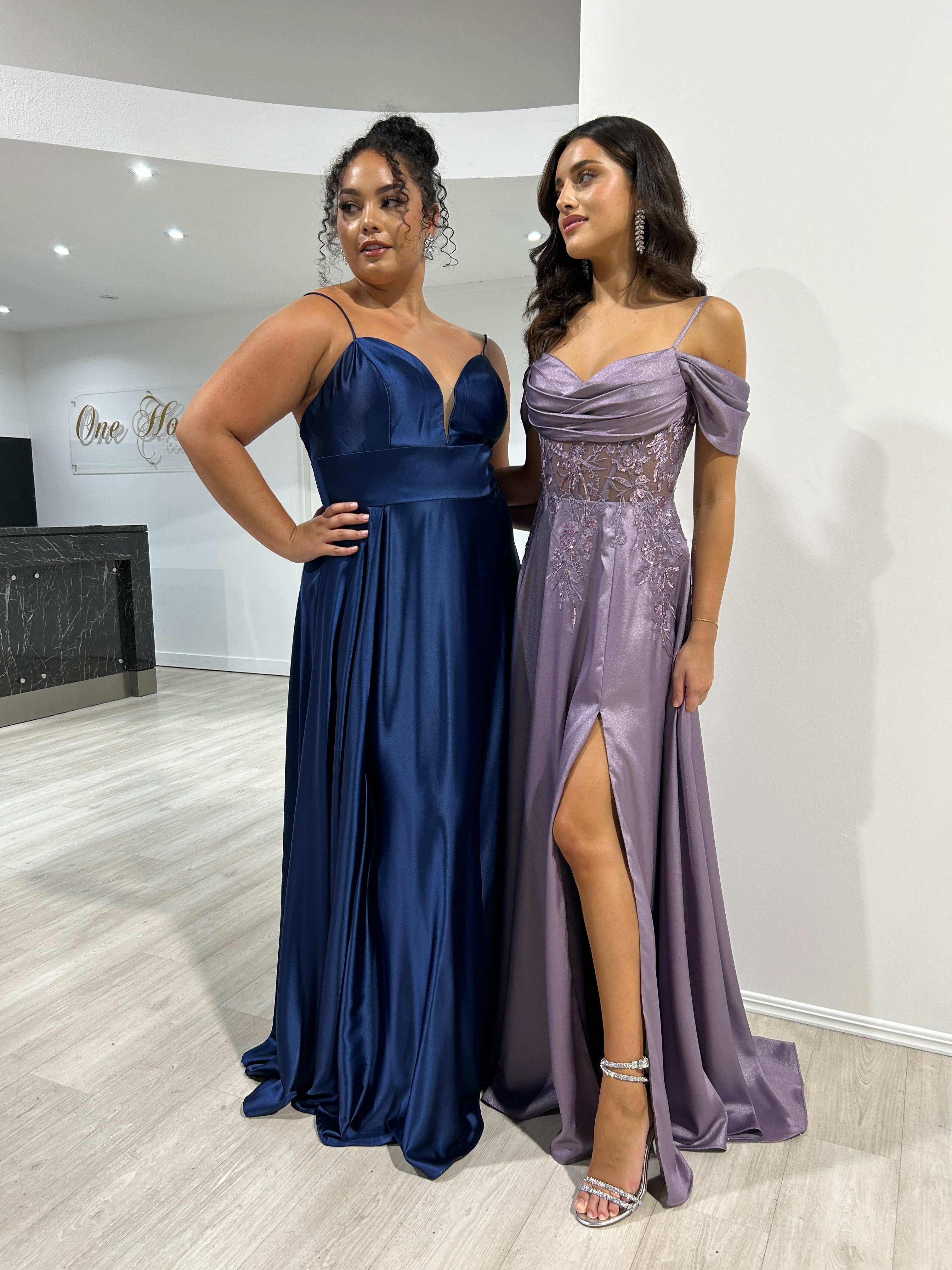 Women in blue and purple satin evening gowns with draped and embellished designs, posing together indoors.
