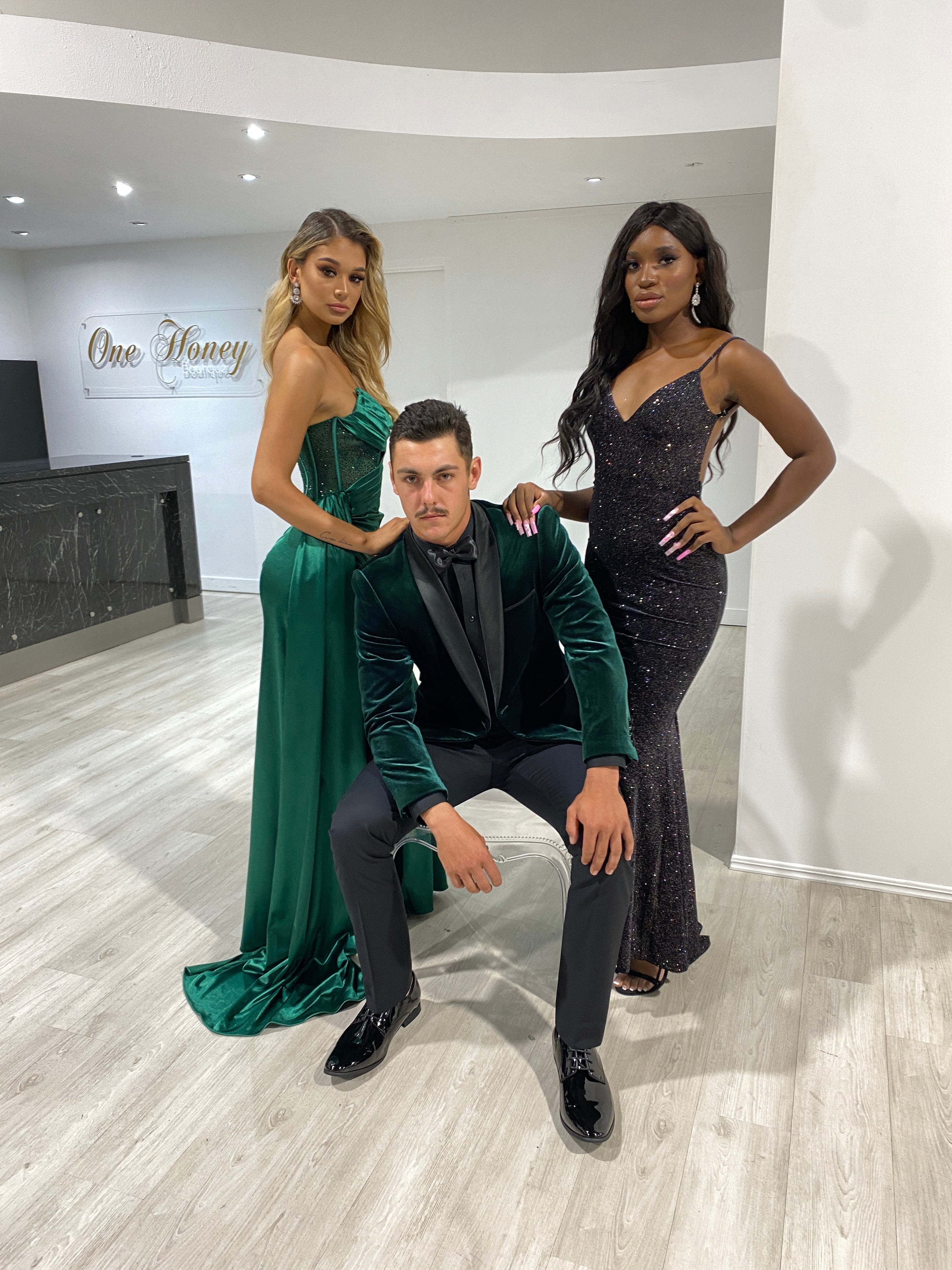 Three models in formal attire, with a woman in a green strapless satin gown, a man in a green velvet suit, and a woman in a black glittery dress.