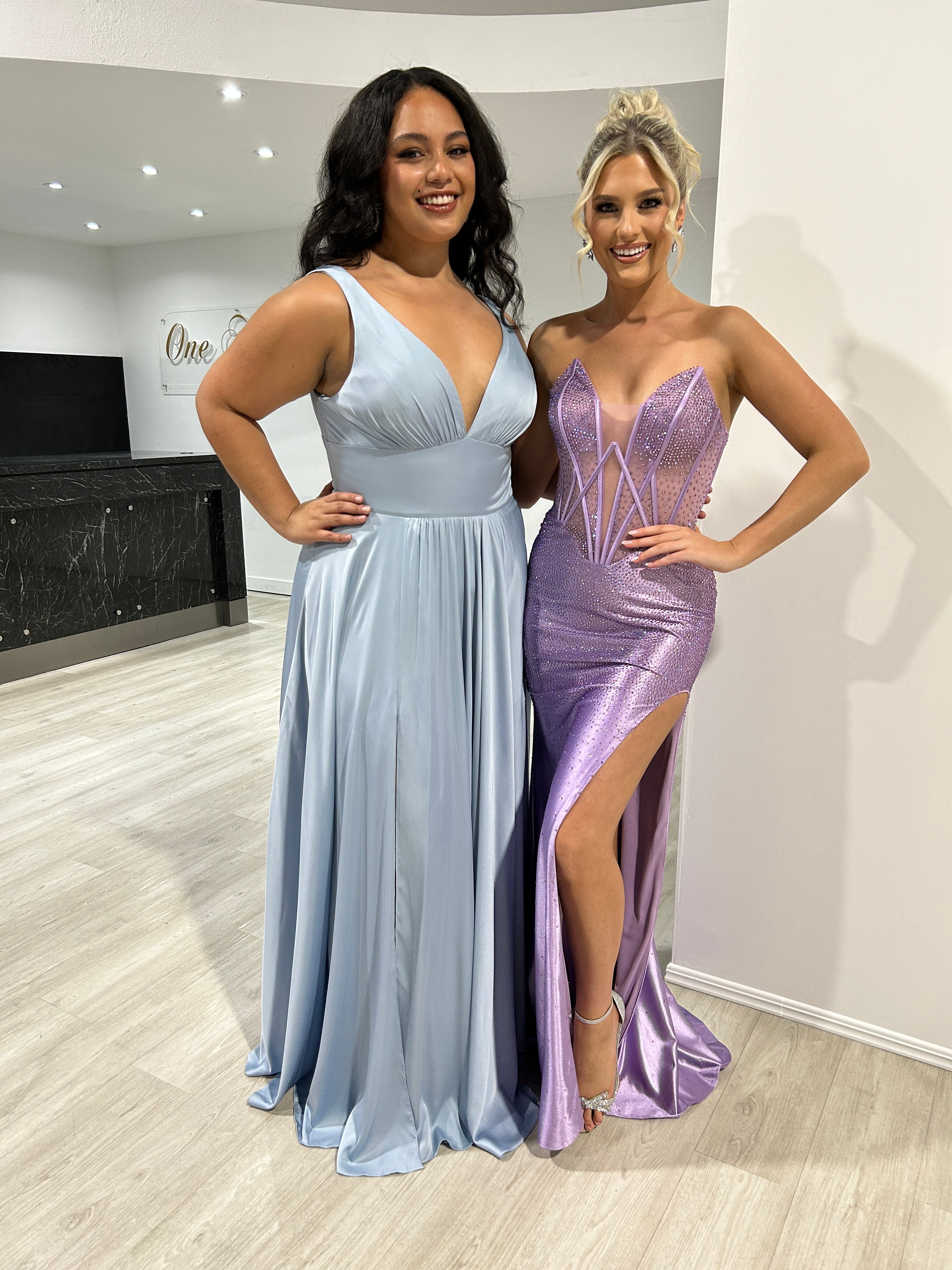Two women posing in formal dresses; the left in a light blue satin gown and the right in a shimmering purple dress with a thigh-high slit.