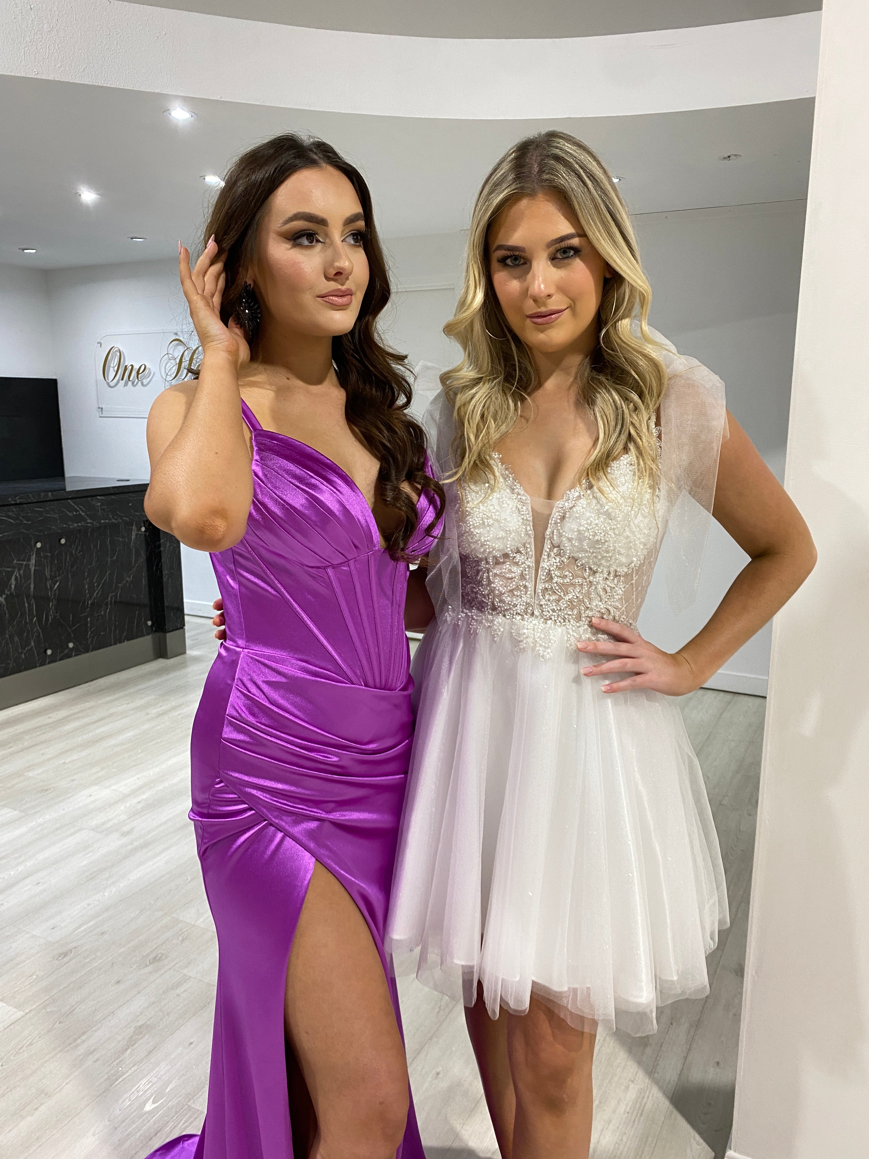 Two women wearing formal dresses; one in a silky purple gown with a leg slit, the other in a white lace mini dress. Brisbane to Gold Coast.