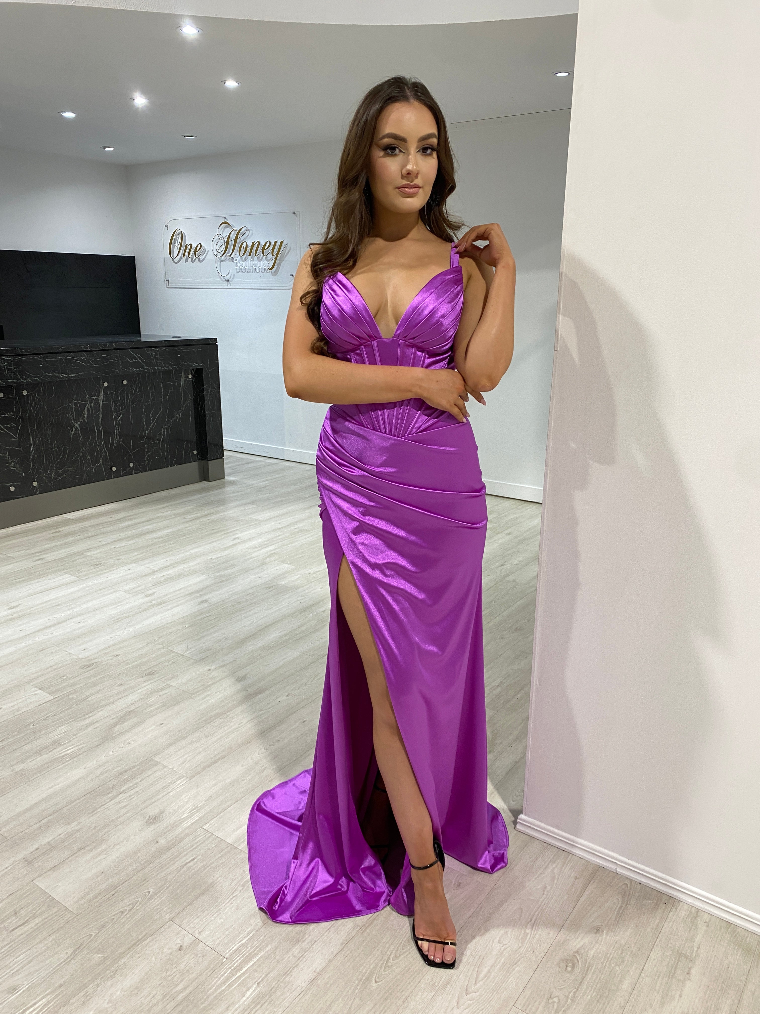 Woman wearing a silk purple evening gown with a deep v-neck and high leg split, posing elegantly indoors, showcasing luxurious style.